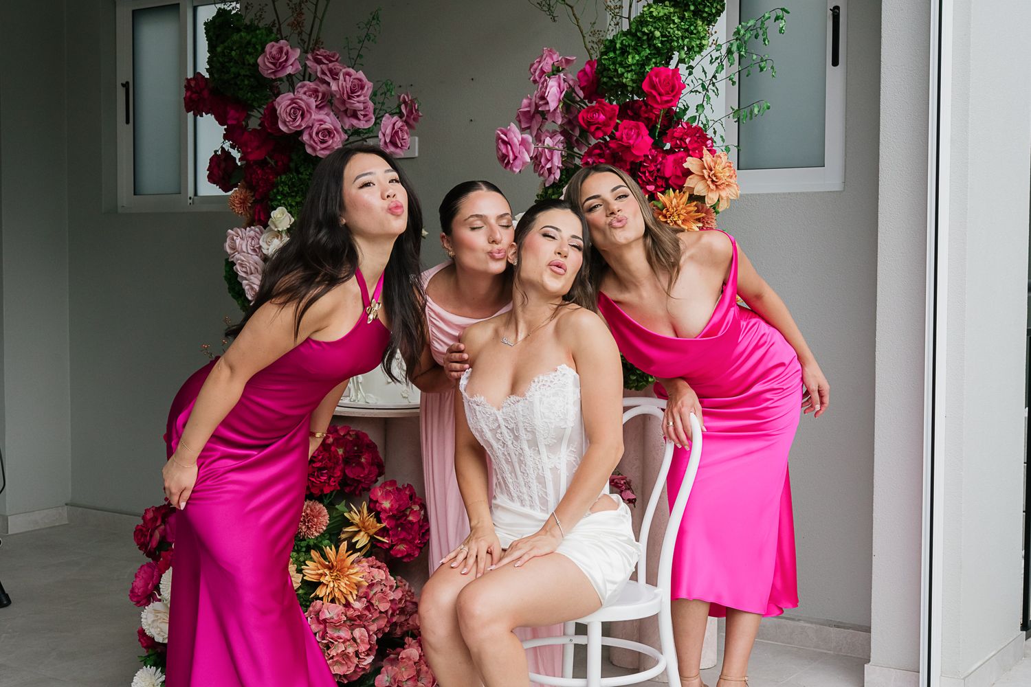 Candid bridal party photo with pink bridesmaid dresses against floral wall installation.