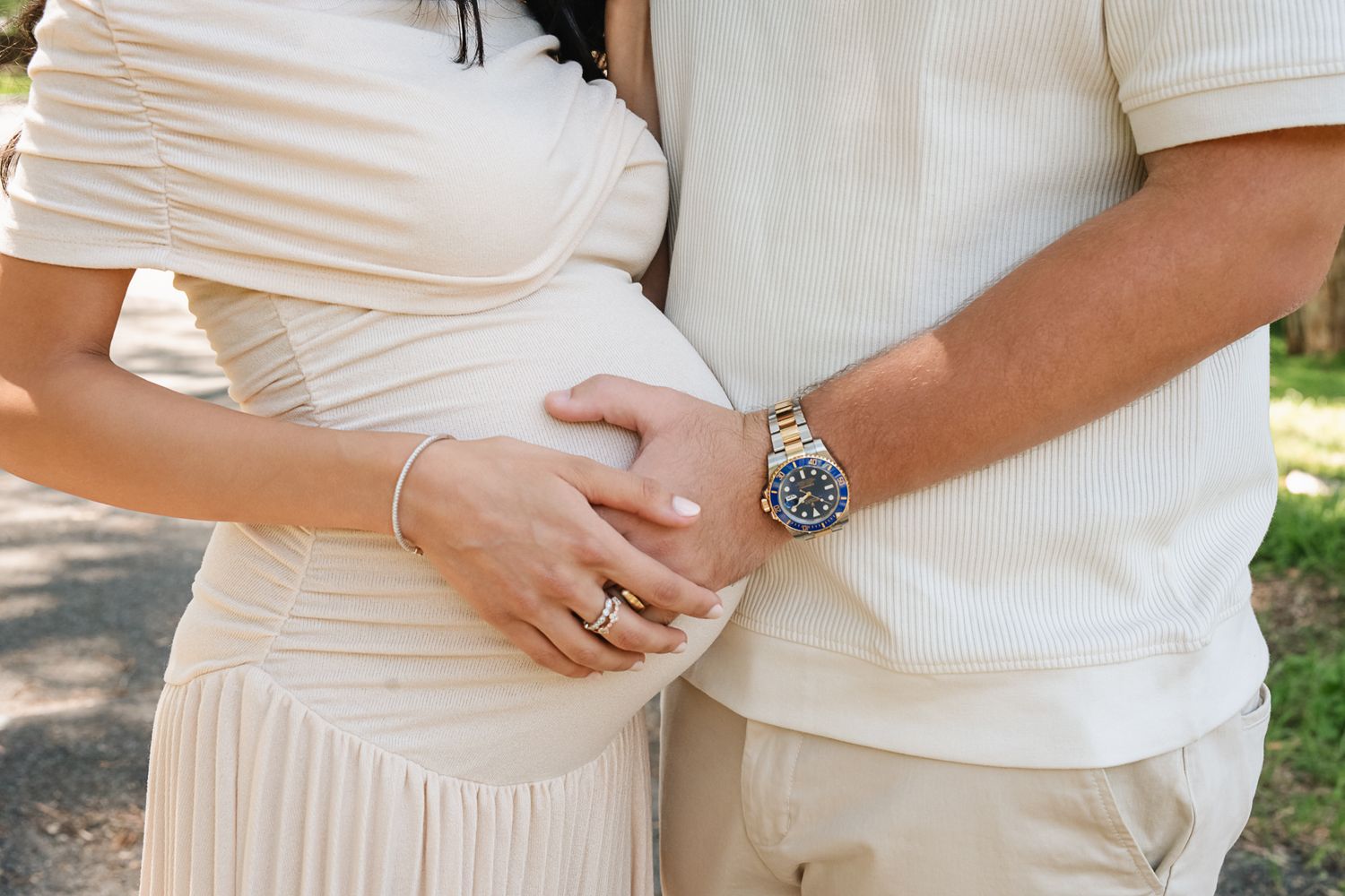 A couple in white outfits embraces while highlighting a baby bump in an outdoor maternity photoshoot.