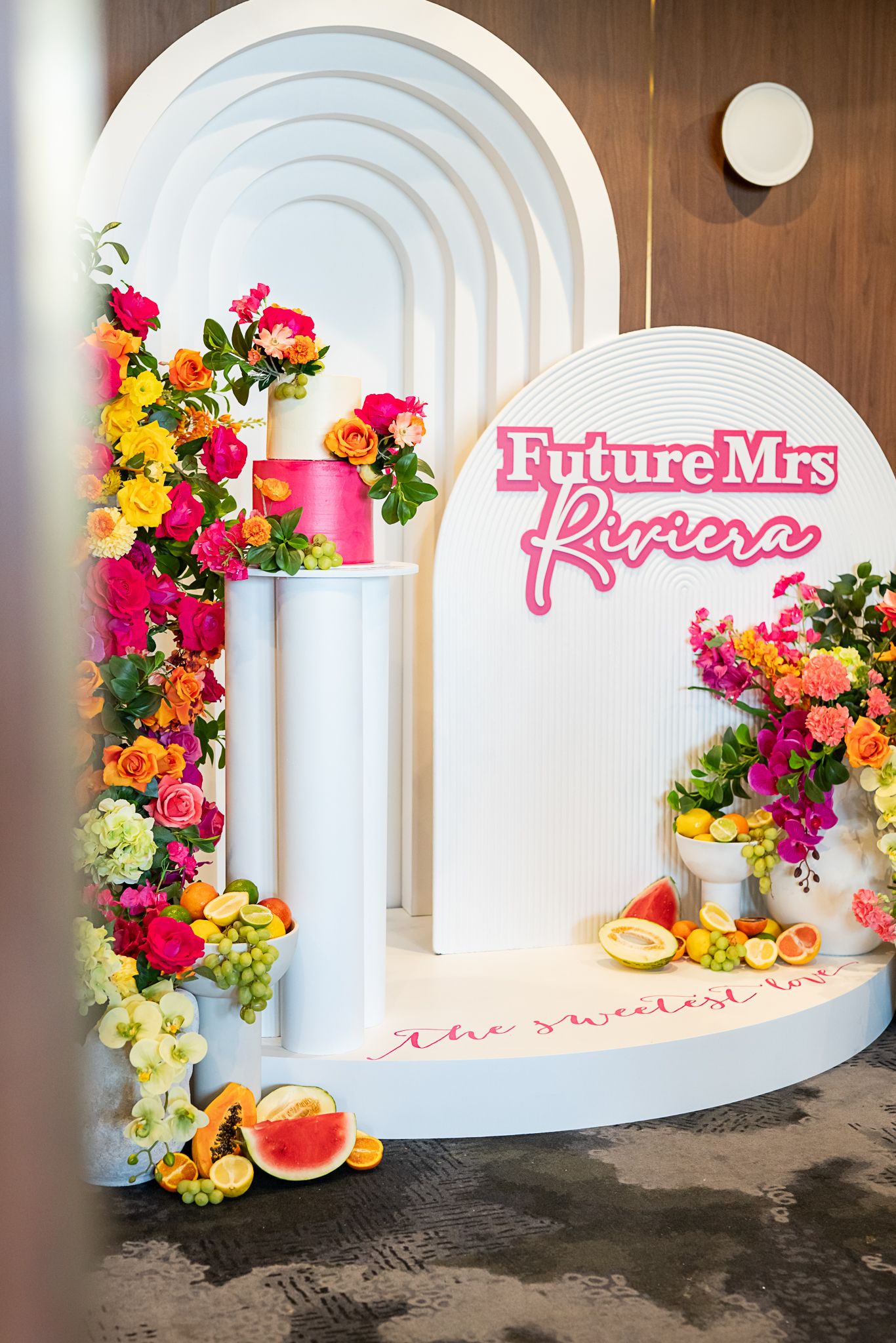 Modern white arch display with pink signage and cascading colorful flowers and citrus accents.