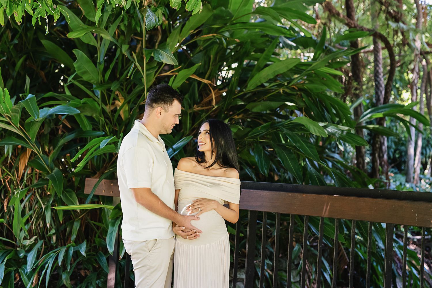 Couple embraces during maternity photoshoot on wooden deck surrounded by tropical plants.
