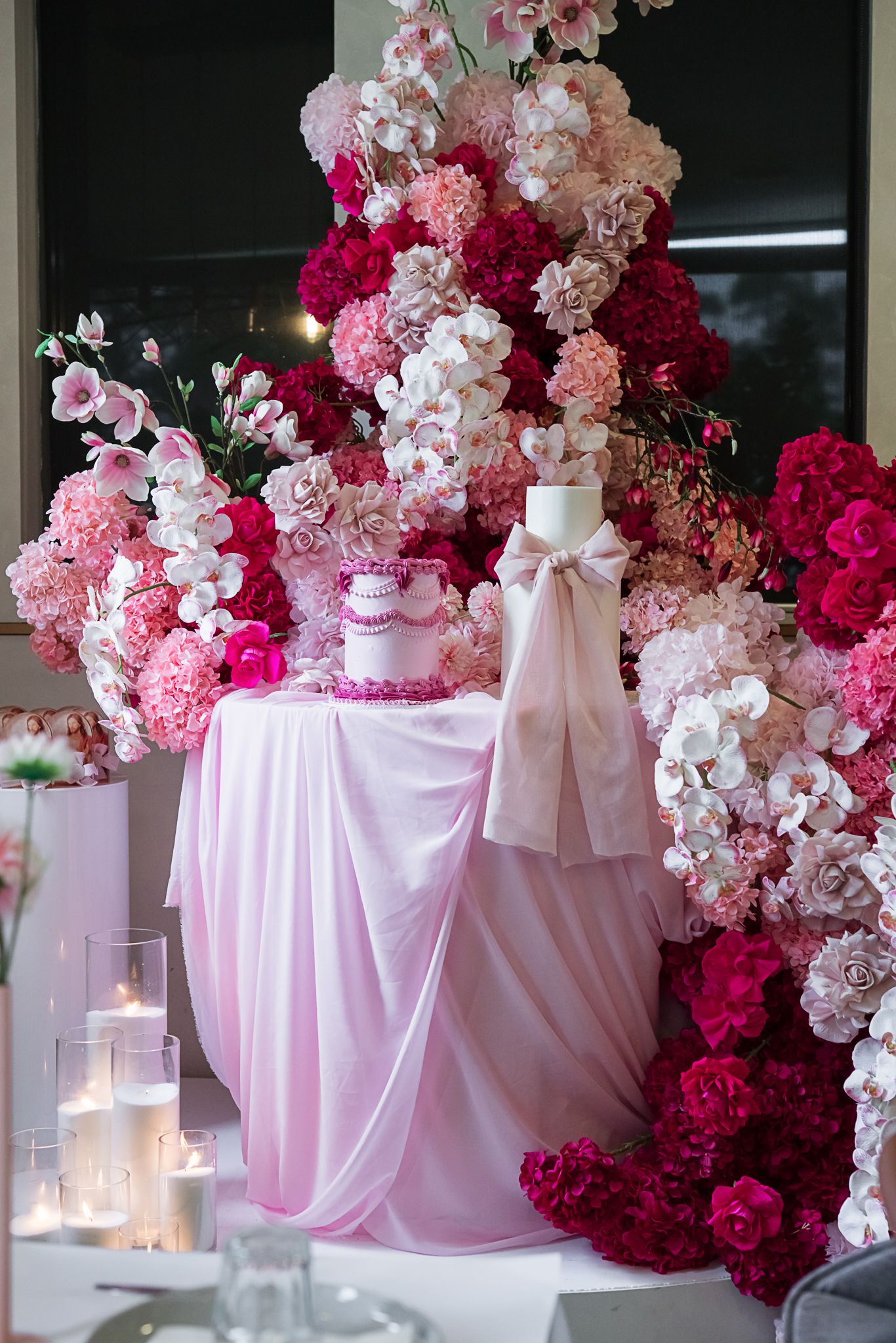 Elegant pink and burgundy floral display with draped table and candles at a wedding dessert table.