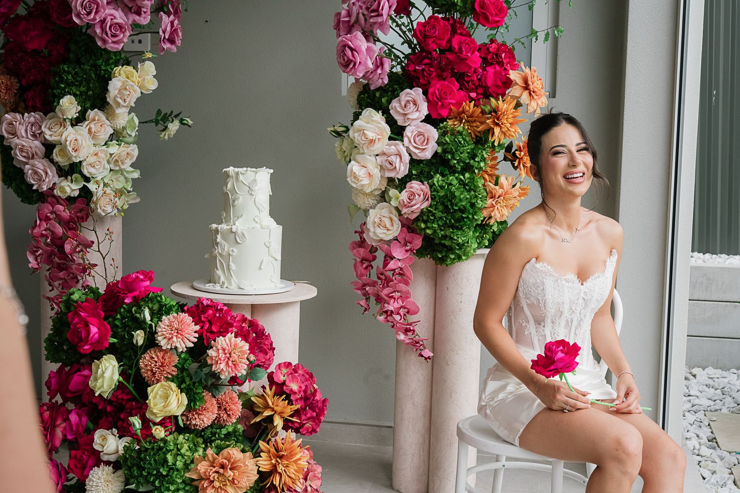 Wedding setting with white tiered cake surrounded by lush pink and red floral arrangements.