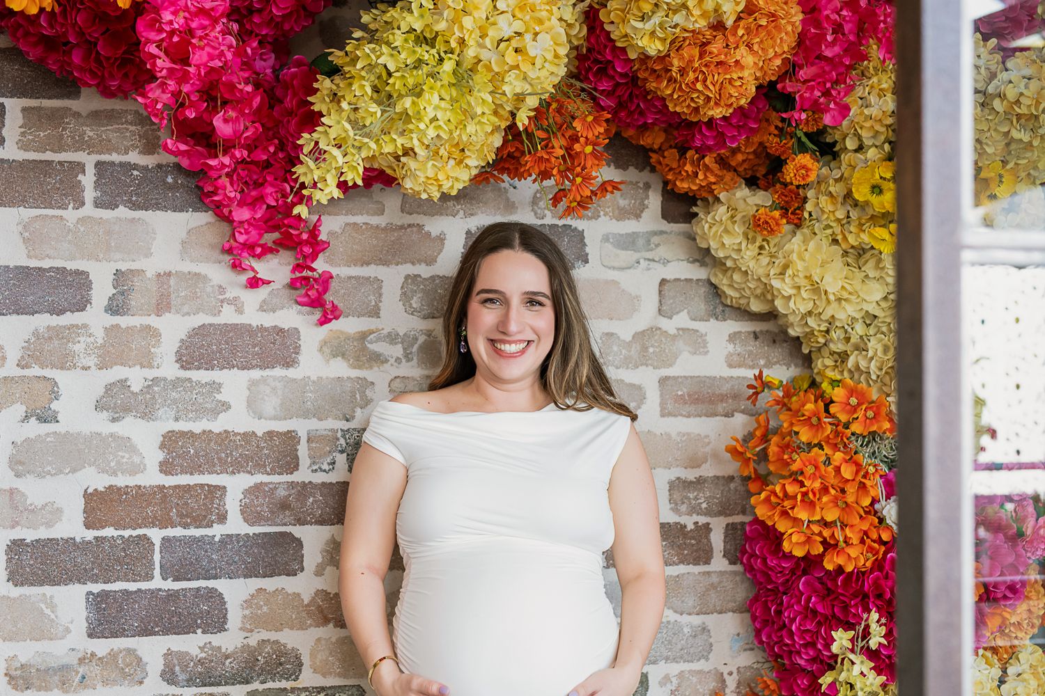 Maternity portrait against brick wall with colorful floral arch installation.