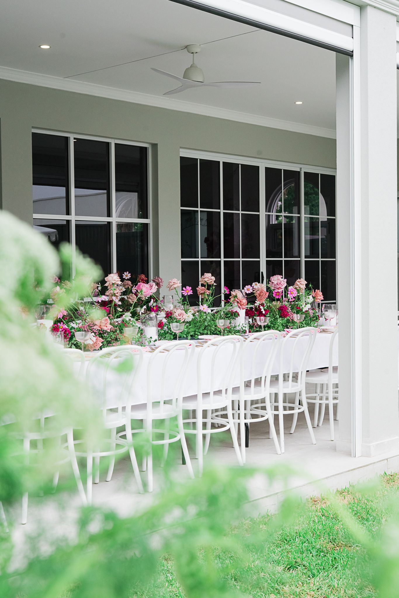 Outdoor dining area with white chairs and pink floral table arrangements viewed through greenery.