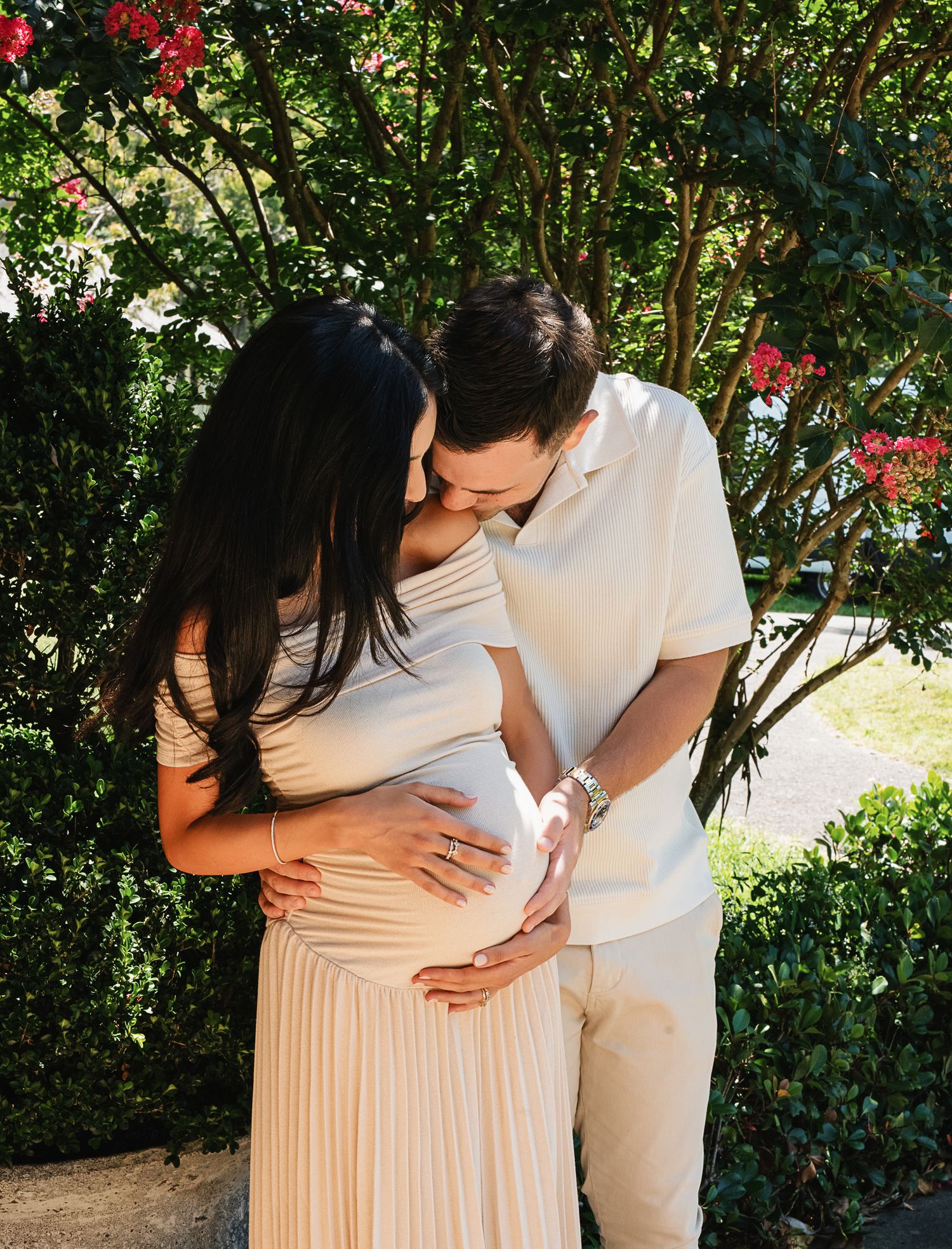 Expecting parents embrace while looking at their baby bump in a romantic outdoor setting.