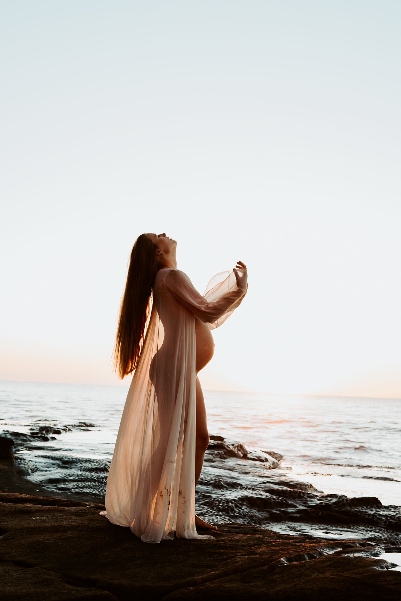Ethereal maternity photo shows figure in flowing dress against sunset ocean backdrop.