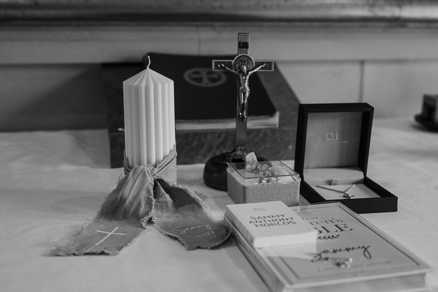 Religious items on white surface including candle, crucifix, rosary beads, and prayer book in black and white photography.