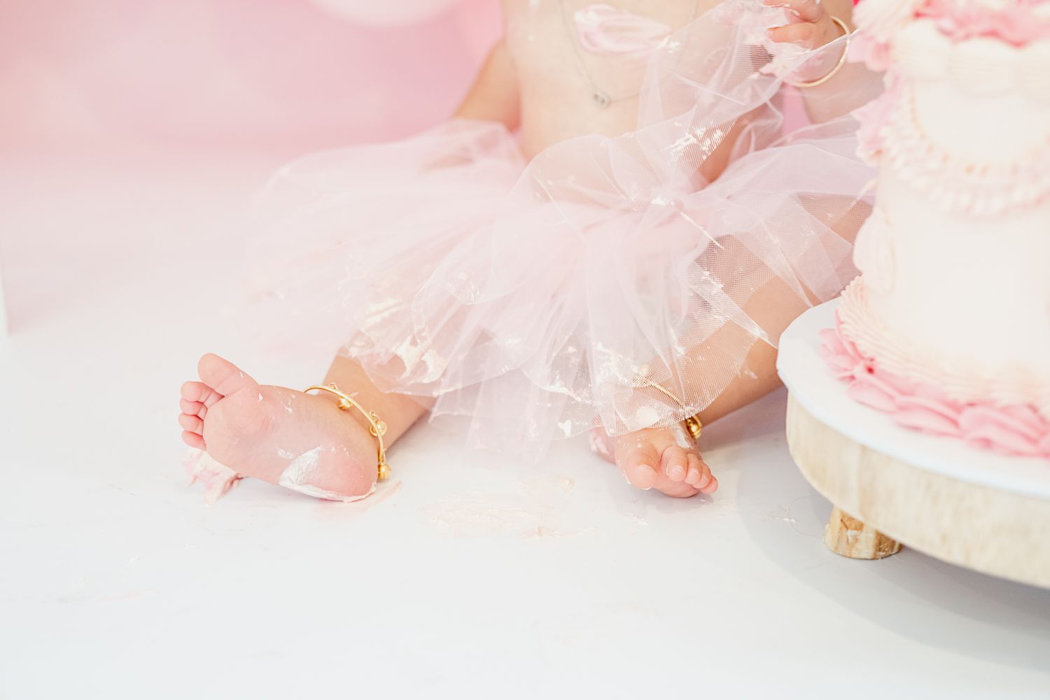 Tiny baby feet peek out from underneath a delicate pink tulle tutu dress near a light colored cake.