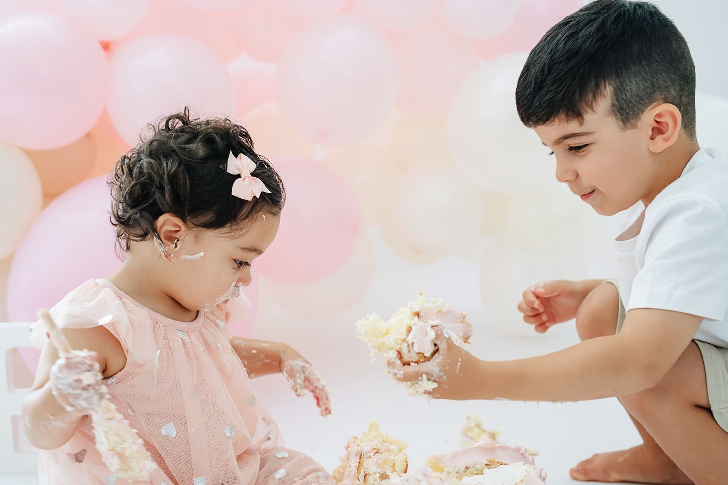 Two children playing with cake during a pink-themed photoshoot with balloons in the background.