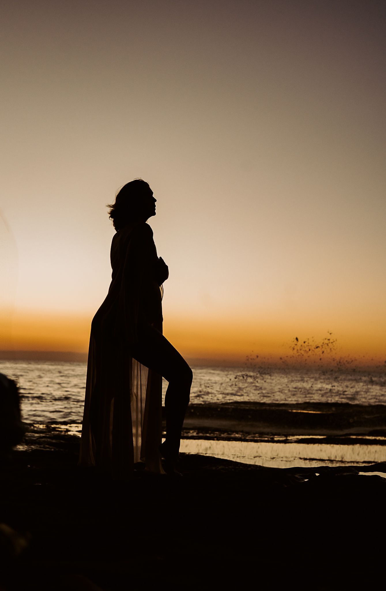 Dramatic silhouette on beach rocks at sunset with flowing fabric in ocean breeze.