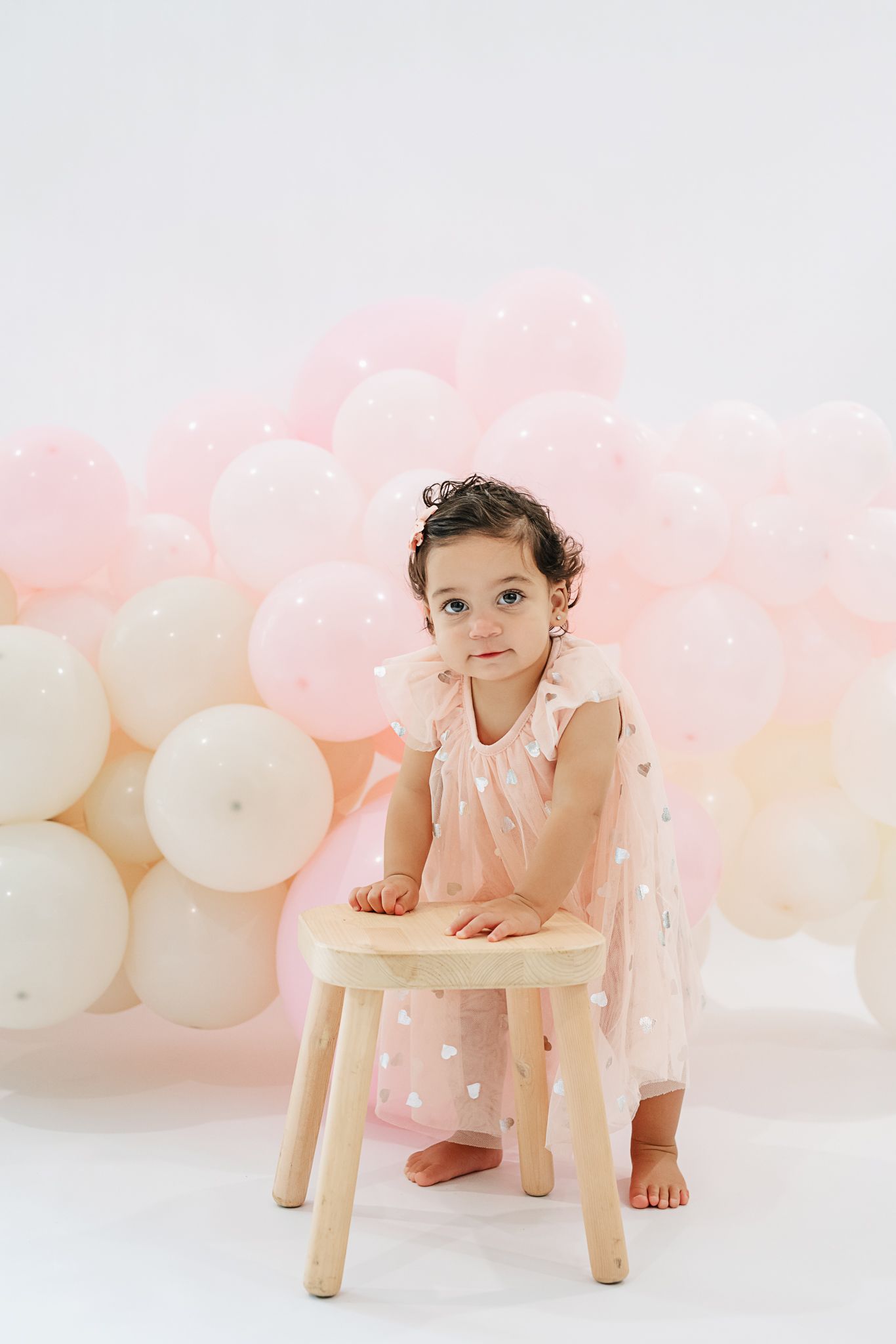 A child in pink dress poses beside wooden stool against pink and white balloon backdrop.