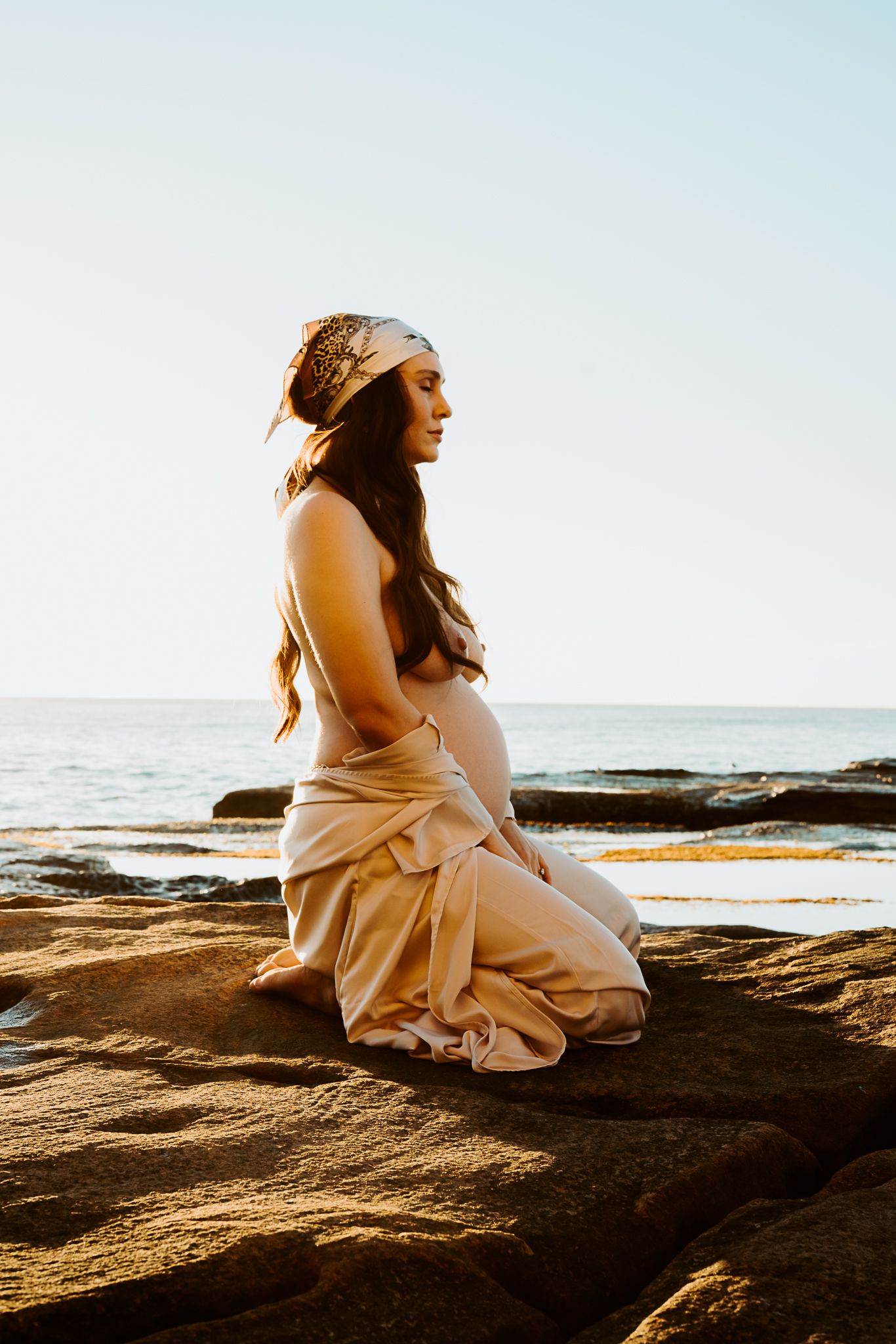 A maternity photo at sunset shows an expectant figure in a flowing beige dress sitting peacefully on coastal rocks.
