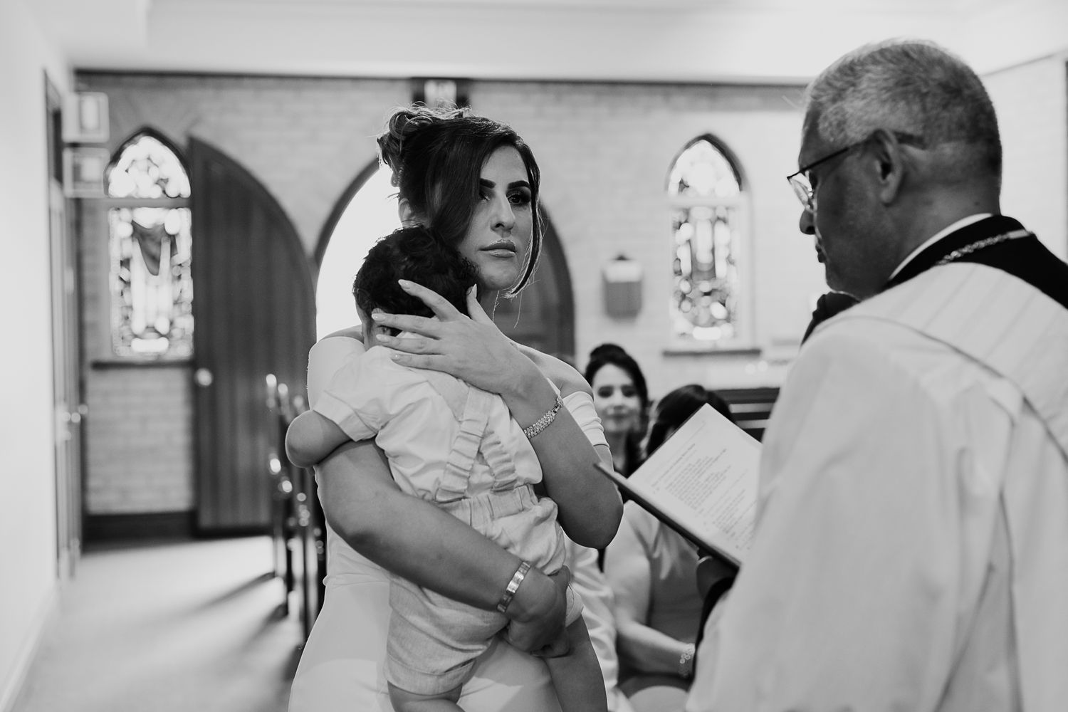 A solemn black and white photograph of a baptism ceremony being performed by a priest in a church.