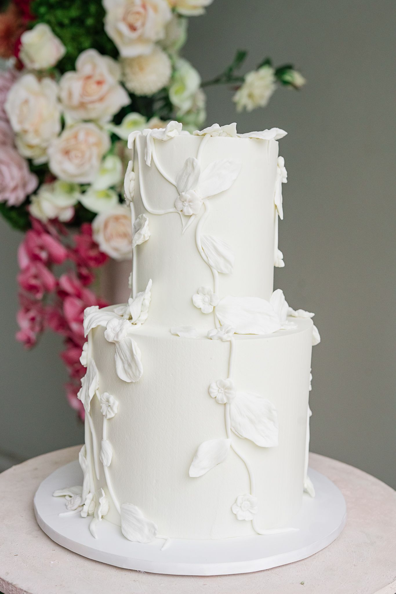 Elegant white two-tier wedding cake with delicate butterfly decorations against floral backdrop.