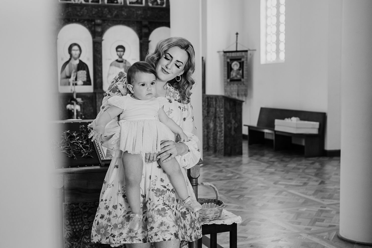 Black and white vintage church interior scene with person in floral dress holding child in baptismal gown.