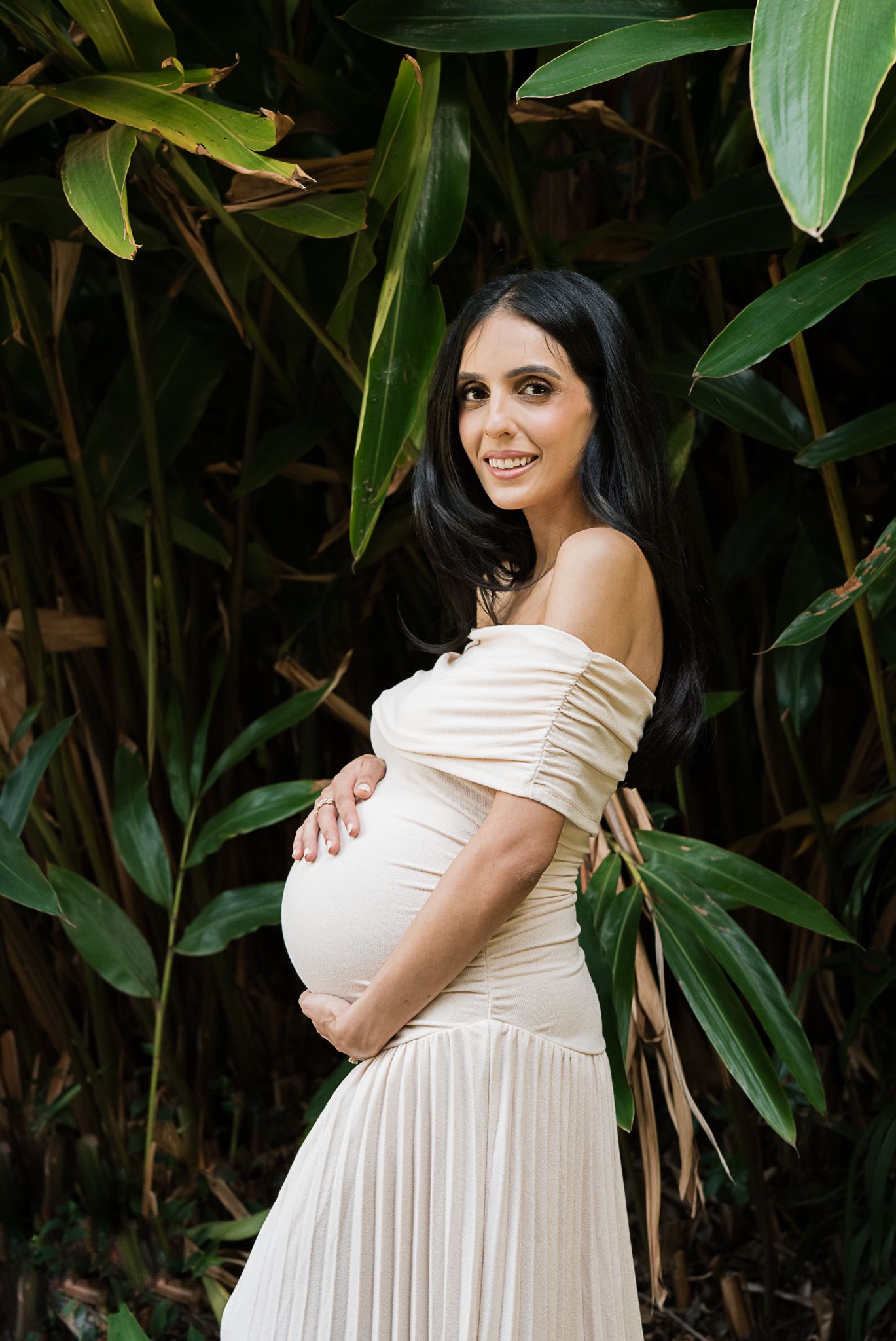 Glowing maternity portrait with cream-colored dress against dark tropical foliage.