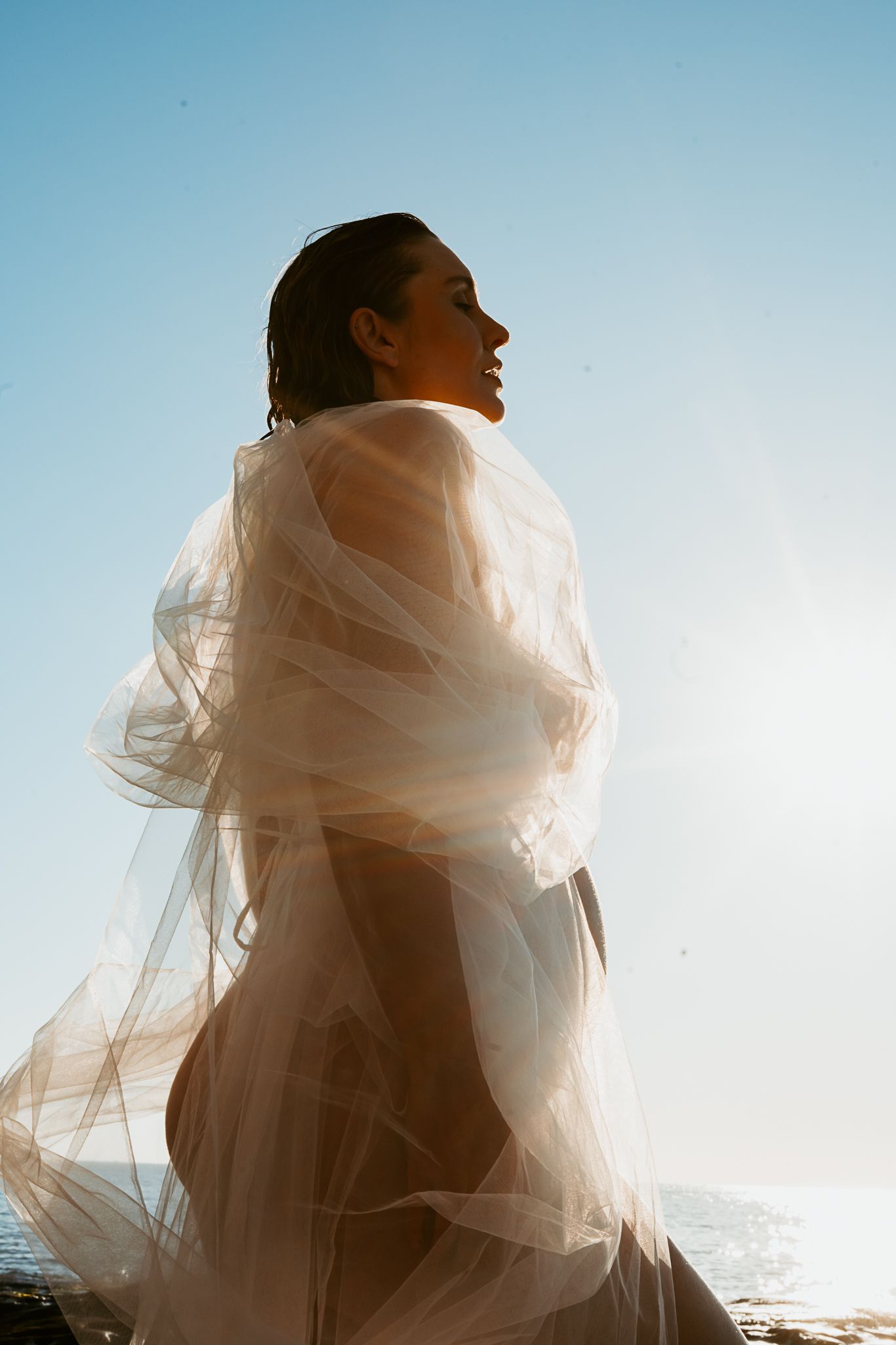 Side profile maternity silhouette wrapped in delicate white fabric against bright sky and ocean backdrop.
