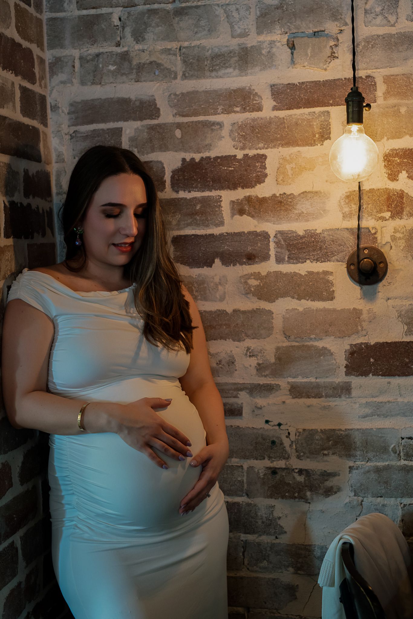 Pregnant woman in white dress poses against rustic brick wall with hanging light fixture.
