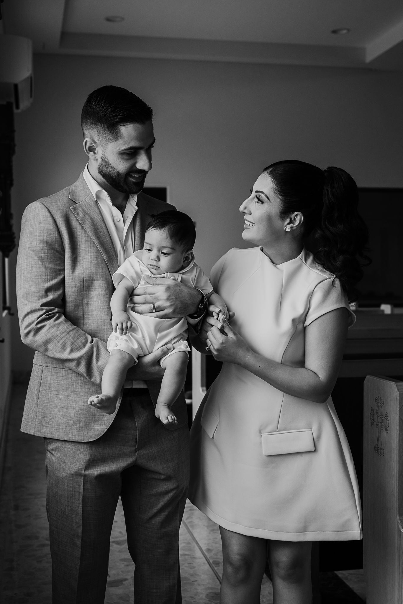 Black and white photo of a couple in formal attire holding a baby while sharing a tender moment indoors.