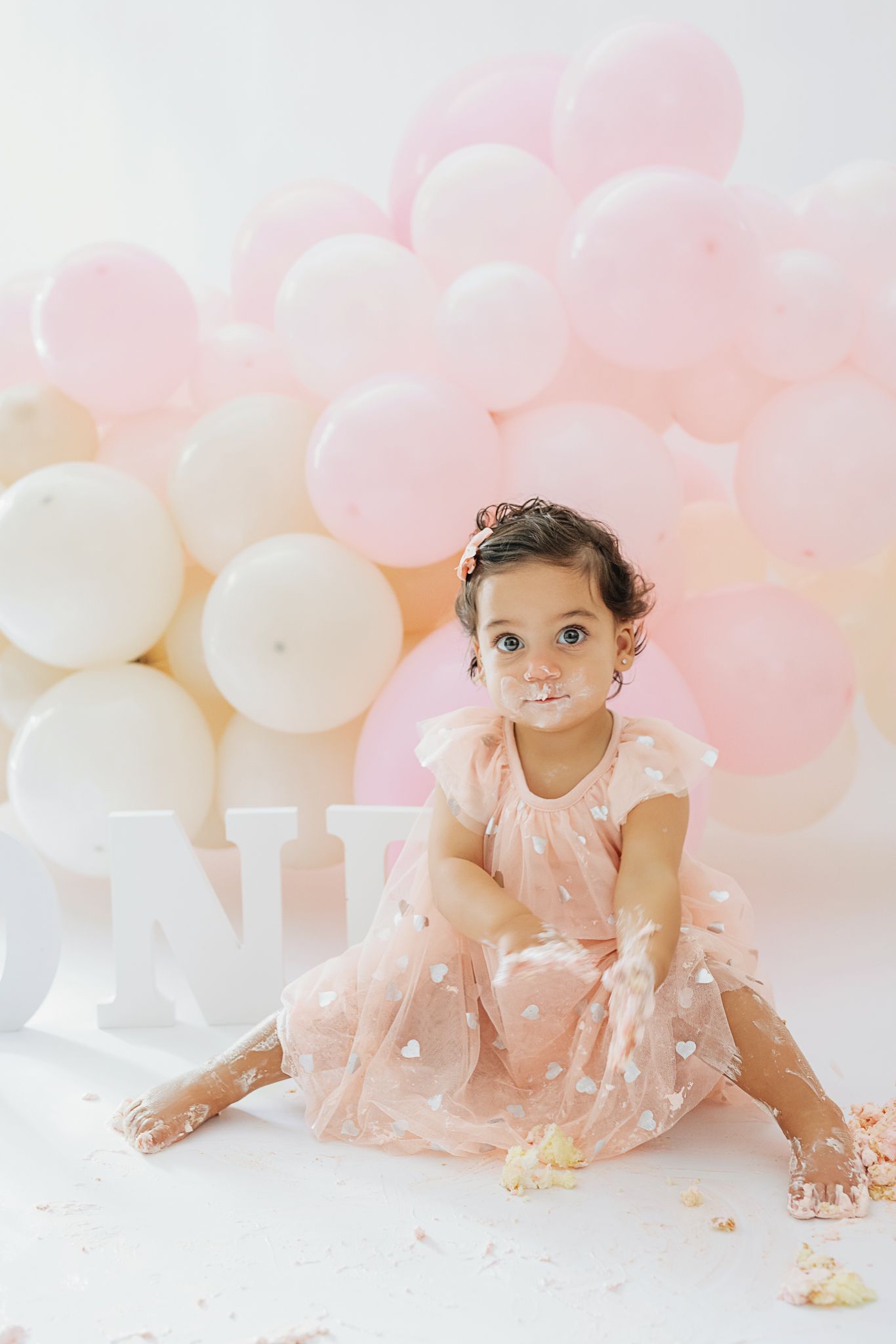 A small child in pink dress sits among pink and white balloons during a photo session.