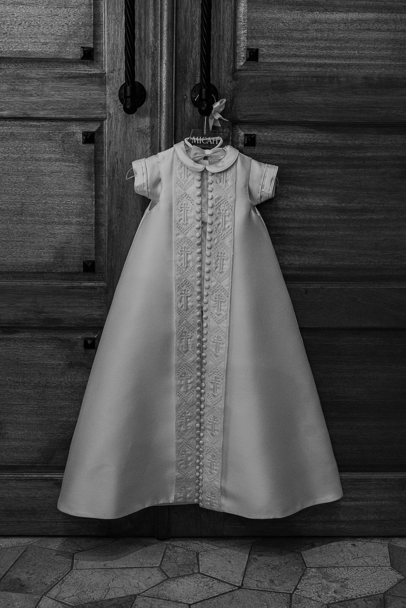 Black and white photo of traditional white christening gown hanging on wooden door.