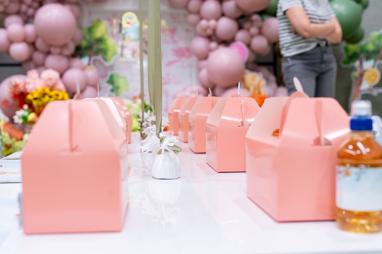 Pink party favor boxes arranged on table with drinks and party decorations.