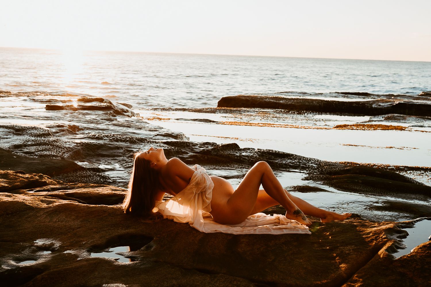 Figure reclining on rocky coastline during golden hour.