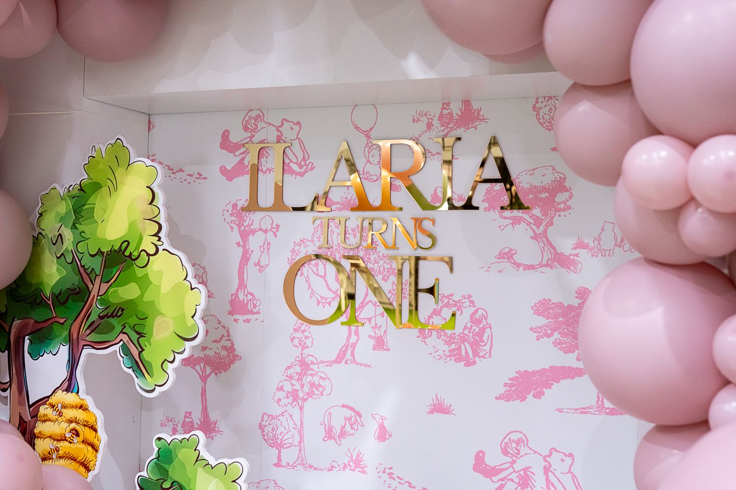 Pink balloon arch frames gold lettering reading 'LARA TURNS ONE' against a decorated backdrop.
