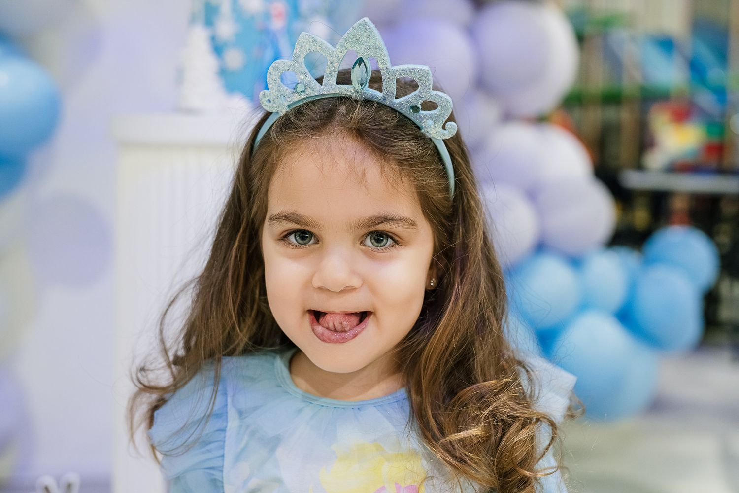 Child wearing a sparkling tiara and blue princess dress smiles at a balloon-decorated party.