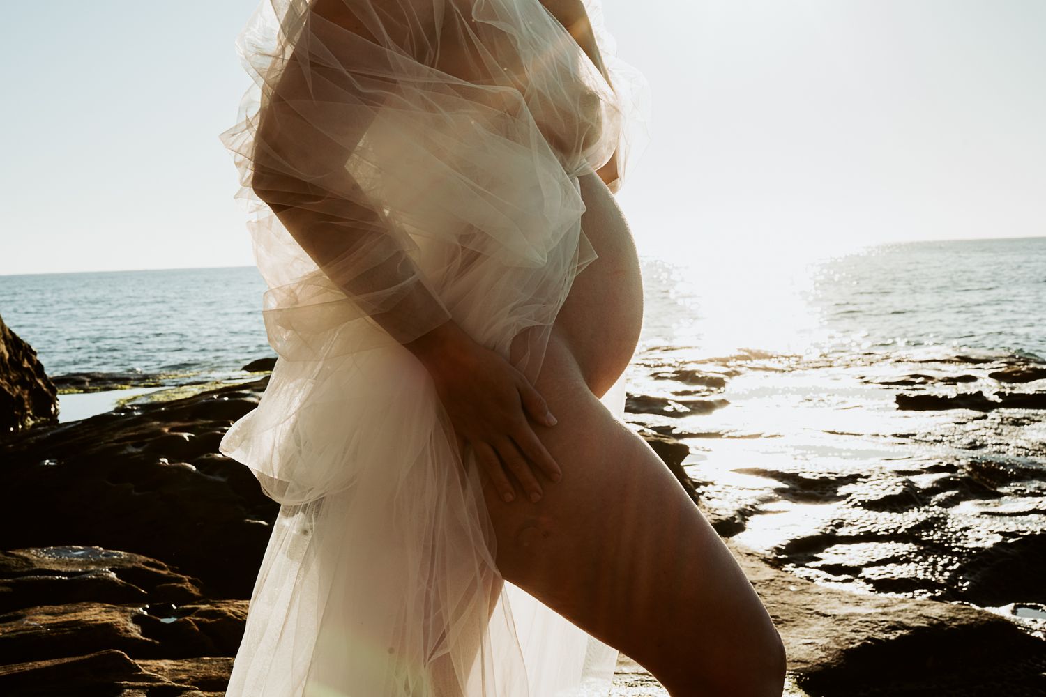 Sun-drenched coastal maternity photo with sheer white fabric draping over rocky shoreline.