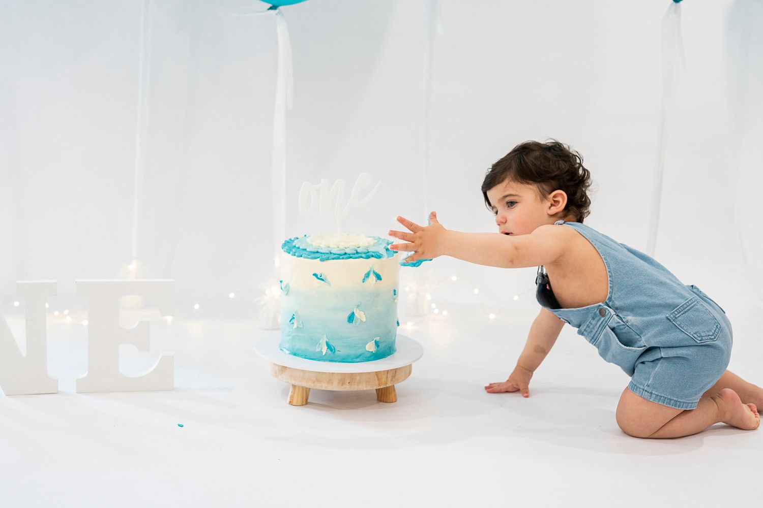 Toddler in blue overalls crawling towards light blue birthday cake on white backdrop.