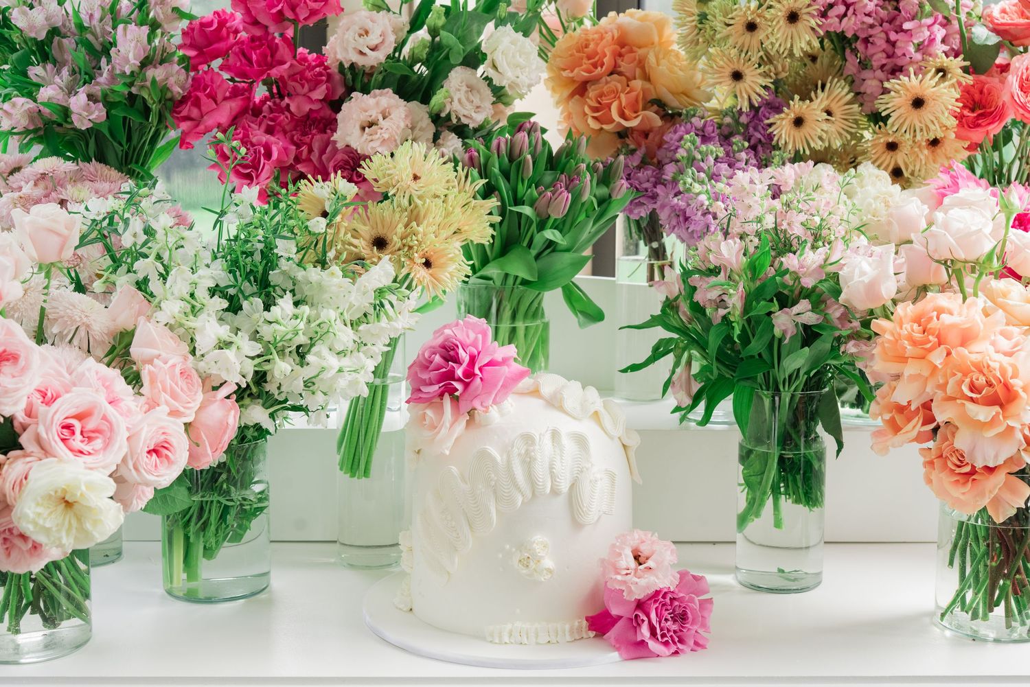 White textured cake surrounded by glass vases filled with colorful roses, carnations and mixed flowers.