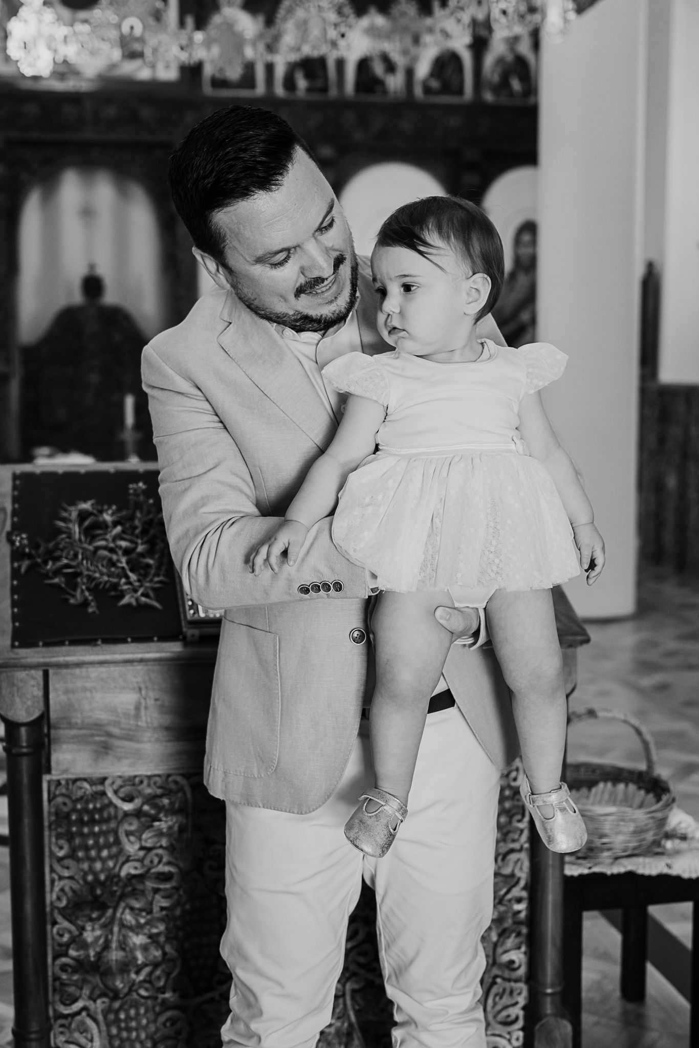 A vintage black and white photograph shows a parent in a white suit lovingly holding a small child in a white dress.