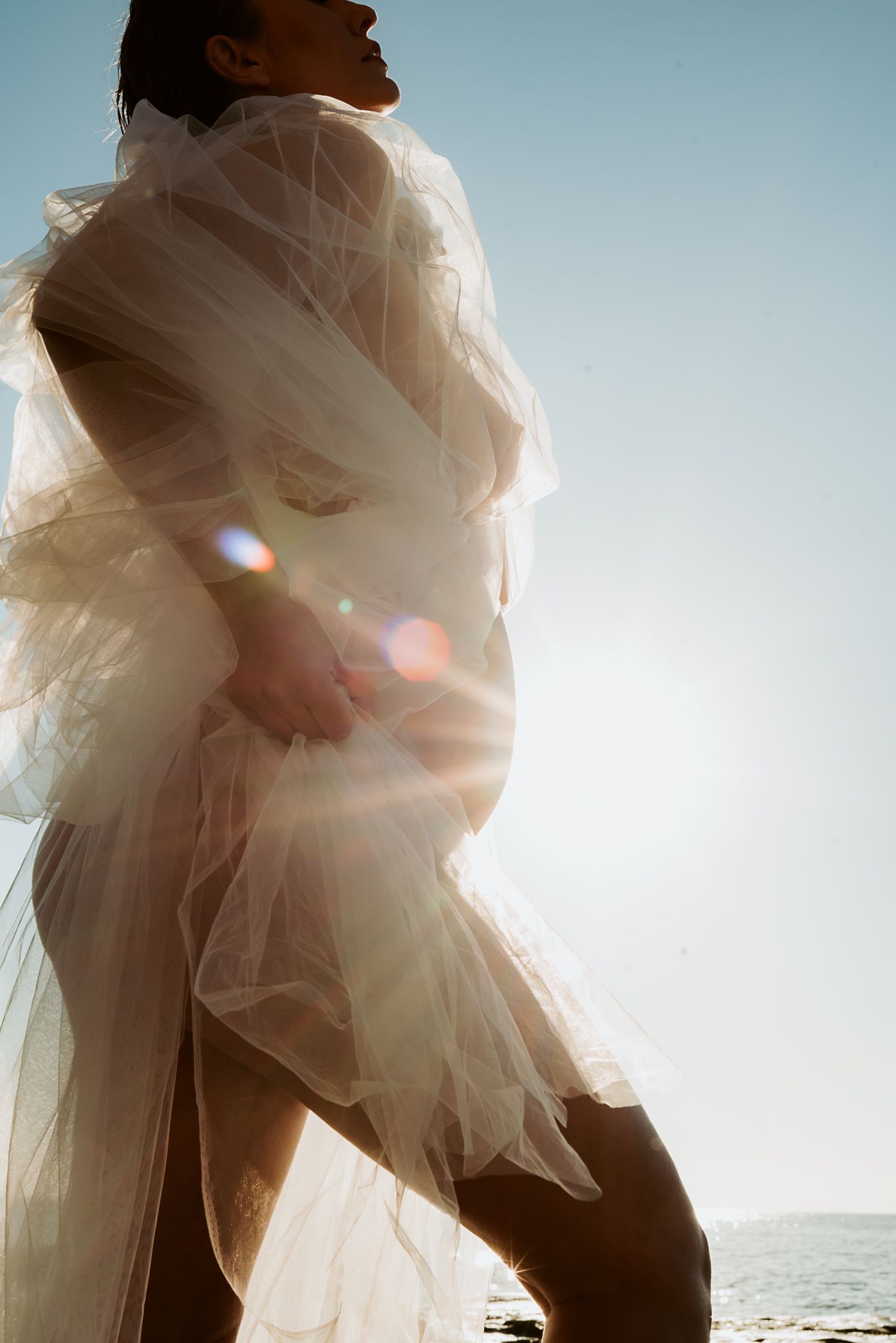Dreamy backlit maternity portrait with sheer fabric flowing in ocean breeze.