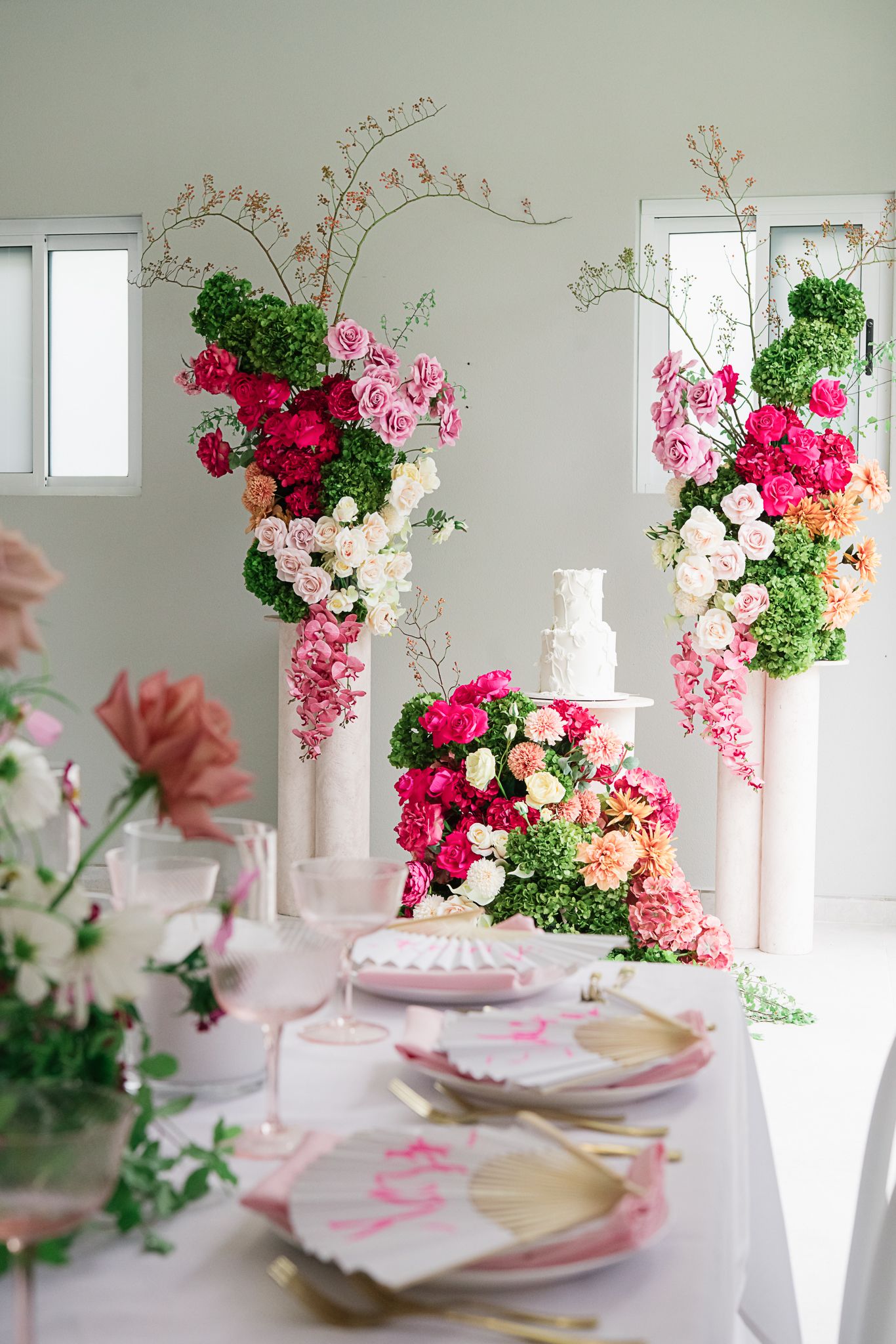 Elegant wedding table setting with dramatic pink floral arrangements and white wedding cake in a bright indoor space.