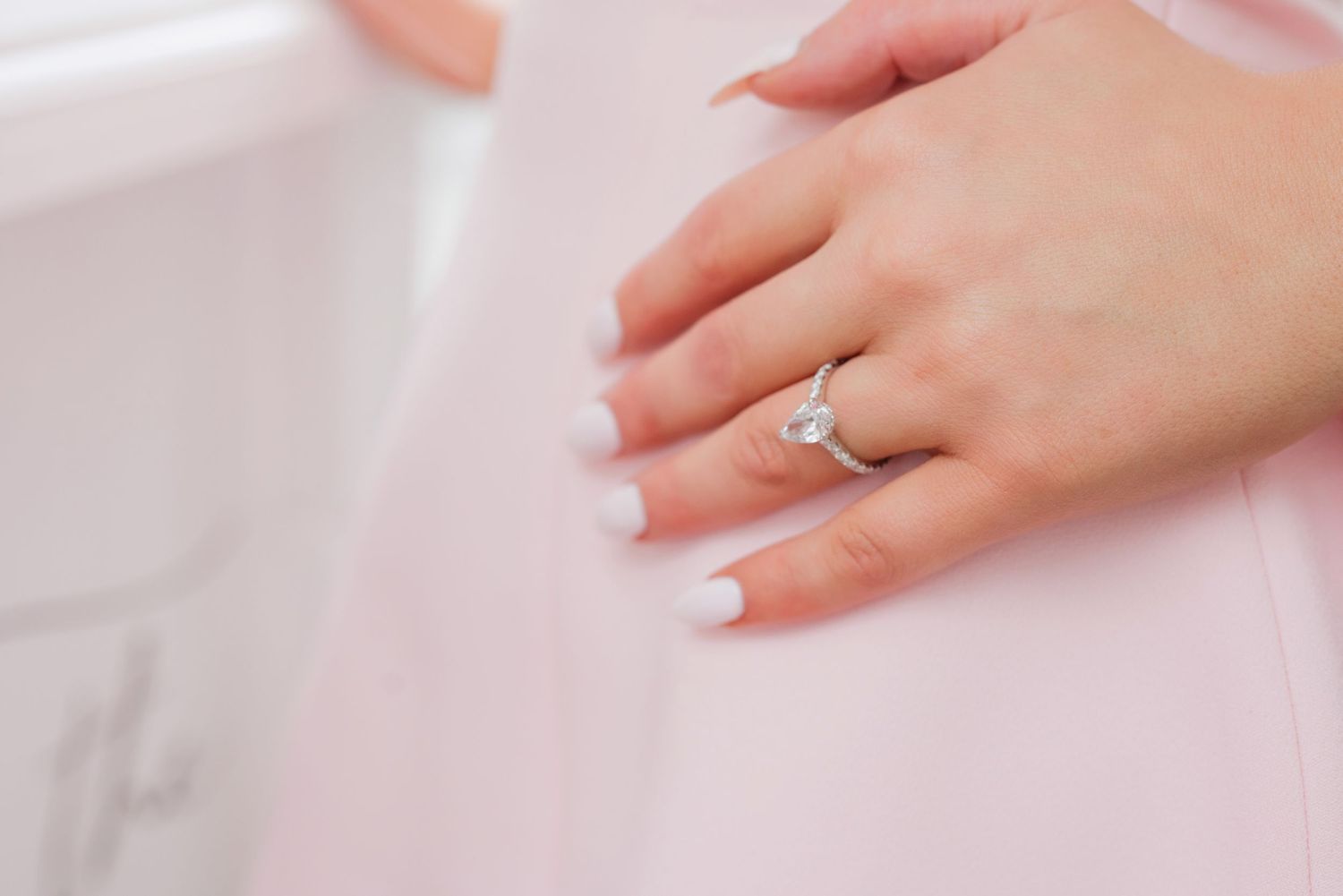 A delicate hand wearing a sparkling diamond engagement ring rests against a soft white fabric background.