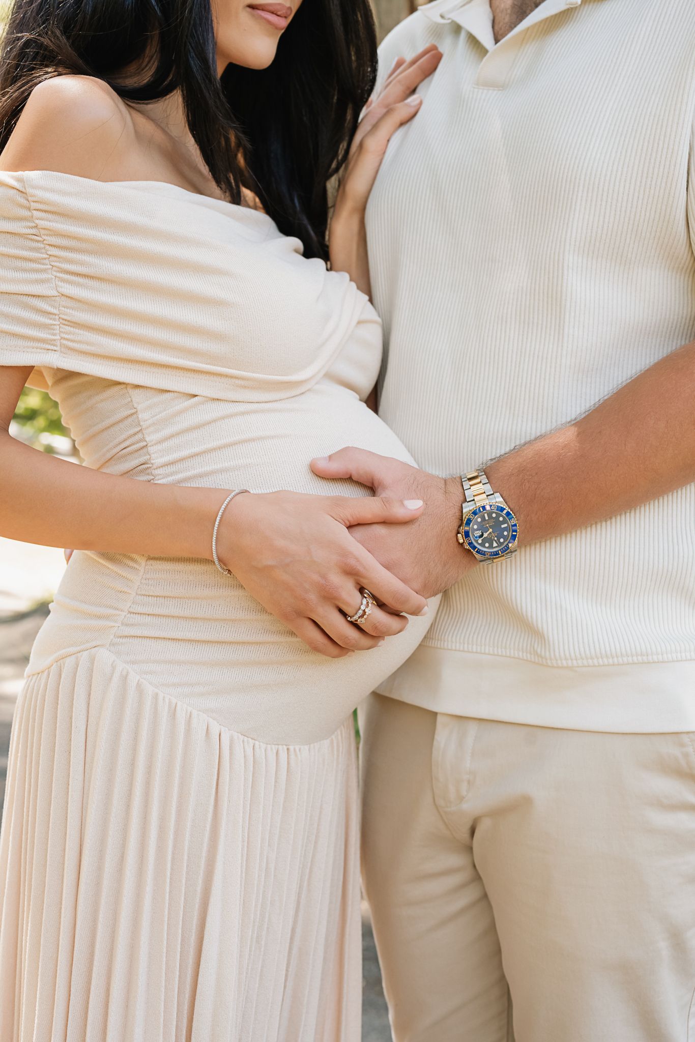 Close-up of expectant parents' hands cradling baby bump while wearing coordinated cream-colored outfits.