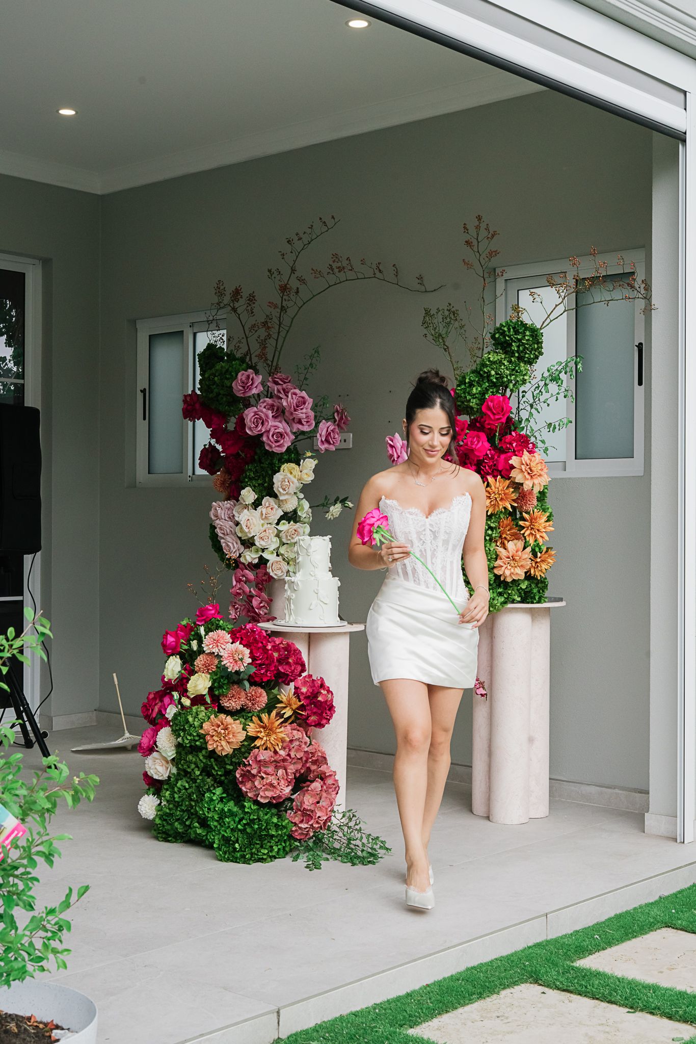 Modern bridal shot in short white dress with dramatic floral installations and outdoor garden setting.