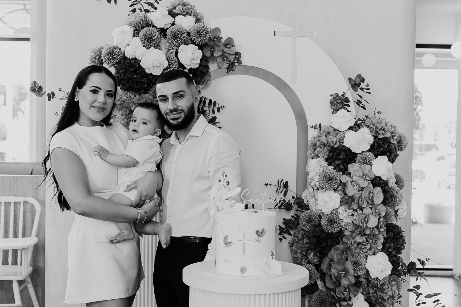 Family poses together next to white wedding cake and floral arch during celebration.