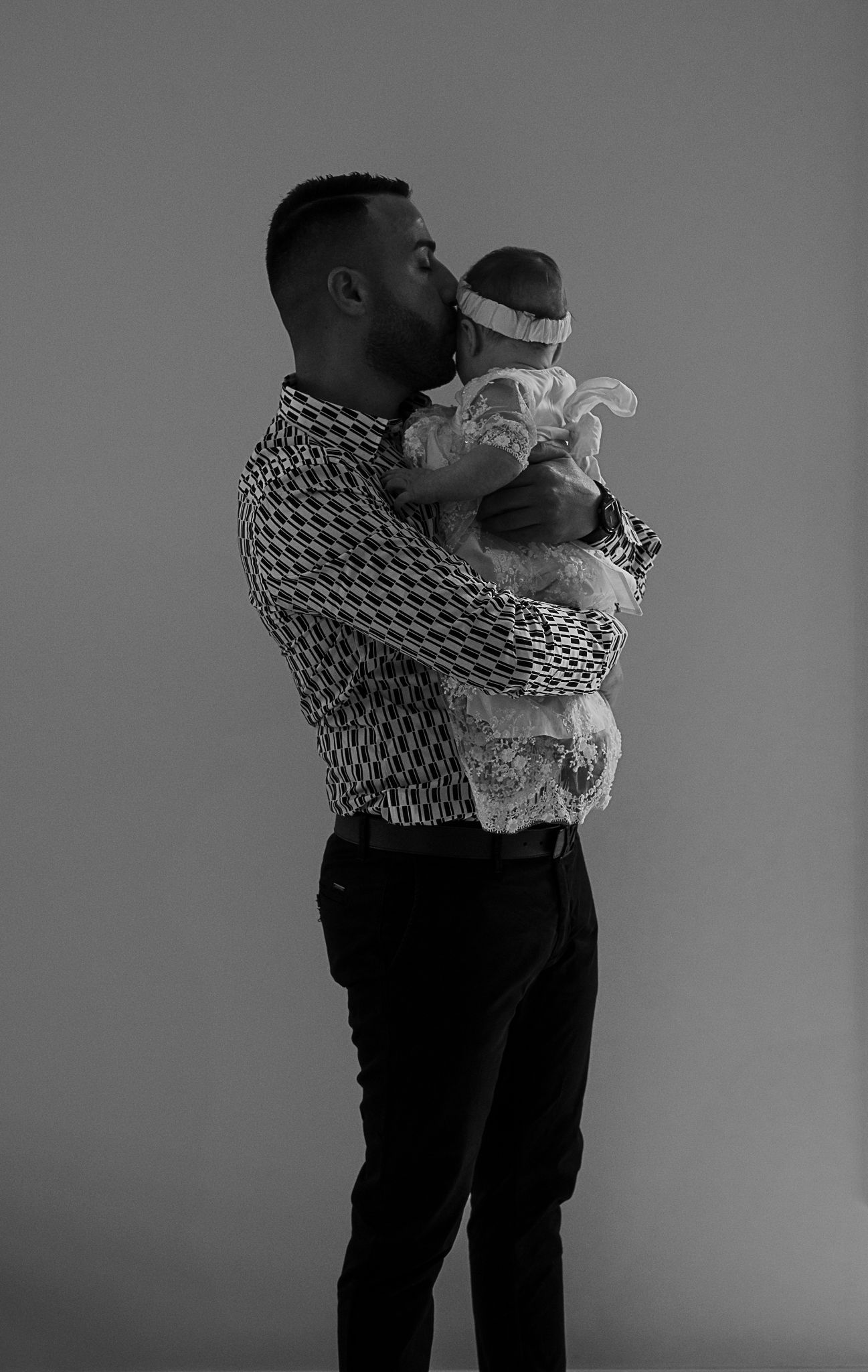 A tender black and white portrait shows an adult in a patterned shirt holding a baby wearing a headband and lace outfit.