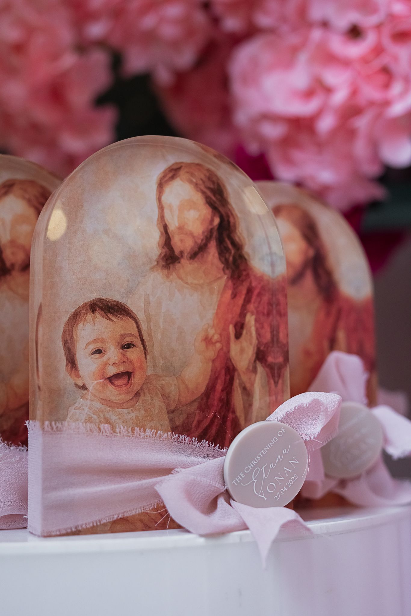 Religious artwork under a glass dome with pink flowers and decorative elements in a soft pastel setting.