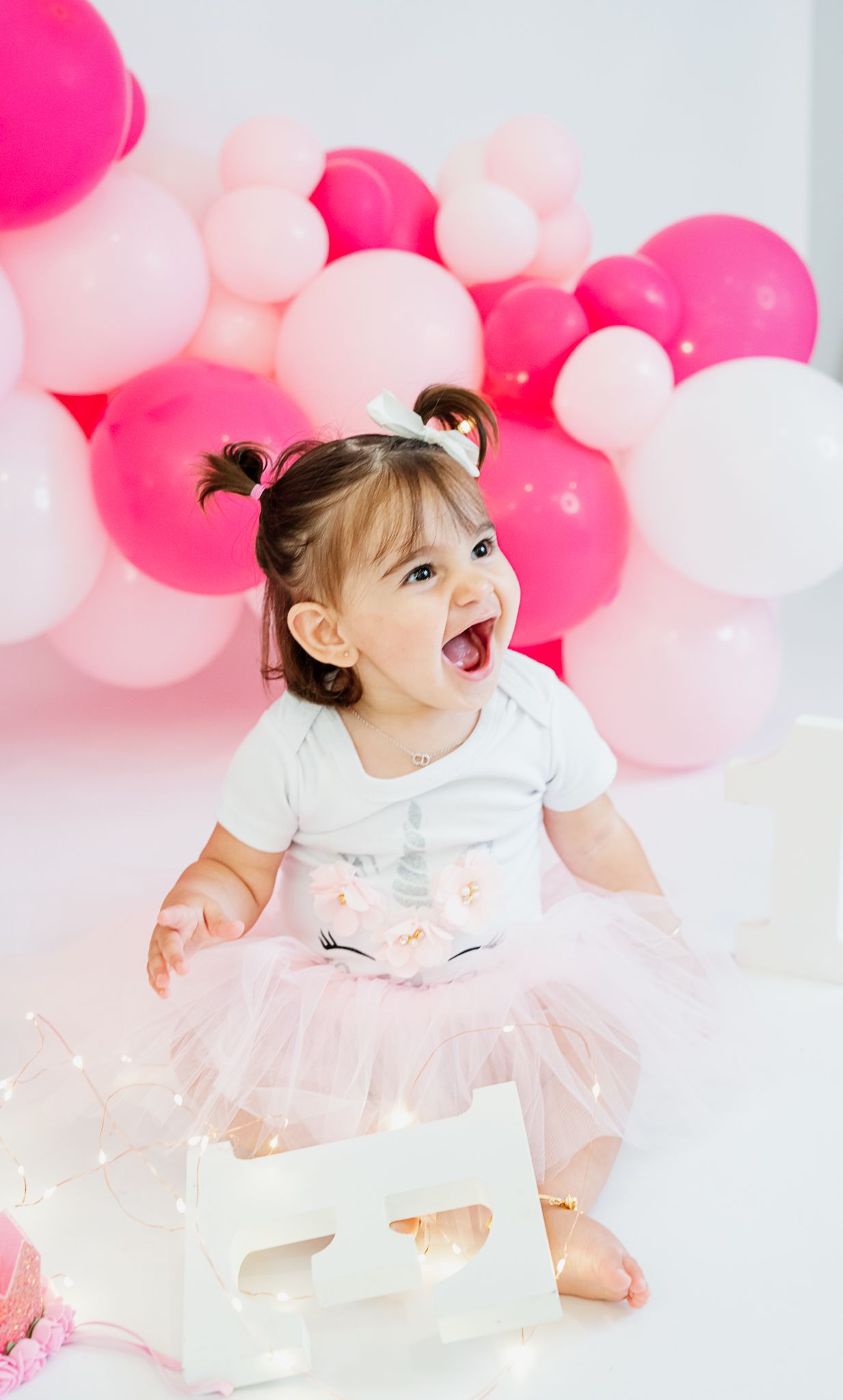 Child in pink tutu celebrates first birthday against pink and white balloon backdrop.