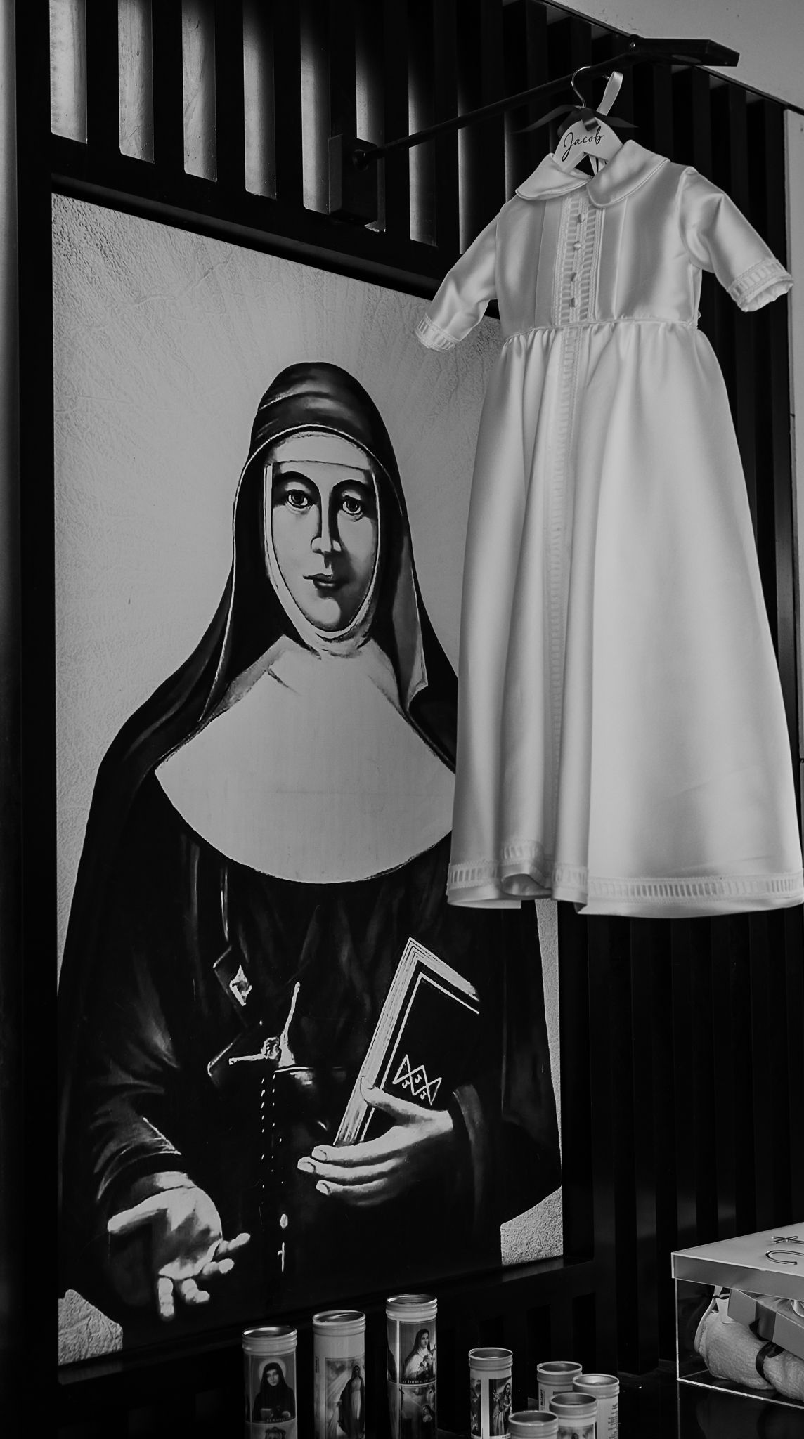 Black and white photo of white baptismal dress hanging next to painting of nun.