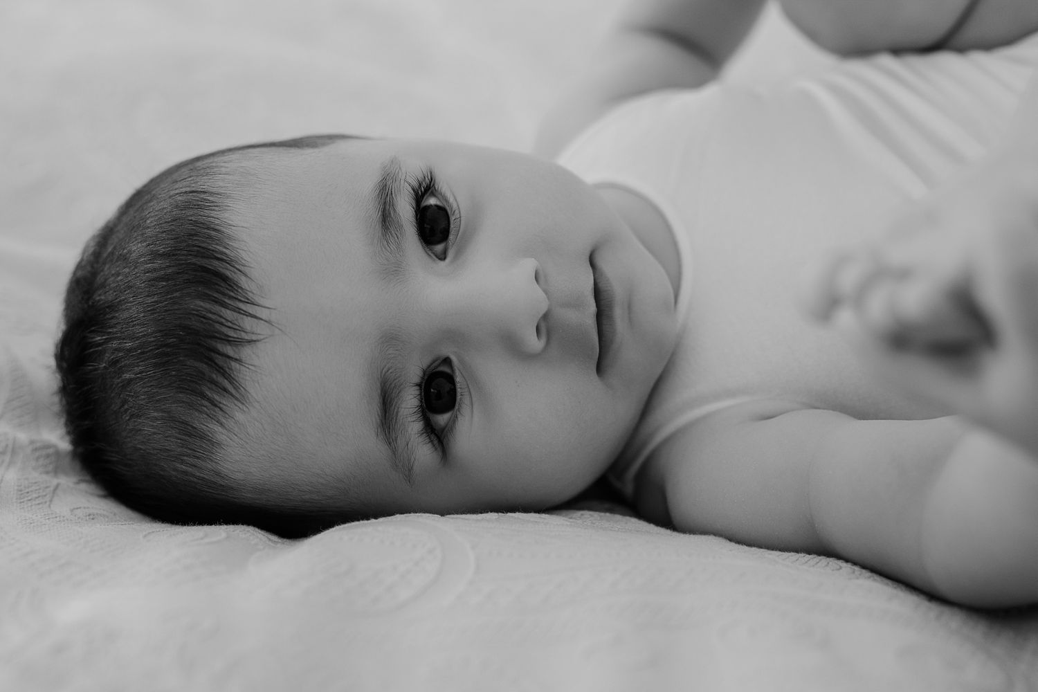 Black and white portrait of a baby lying on soft bedding while looking at the camera.