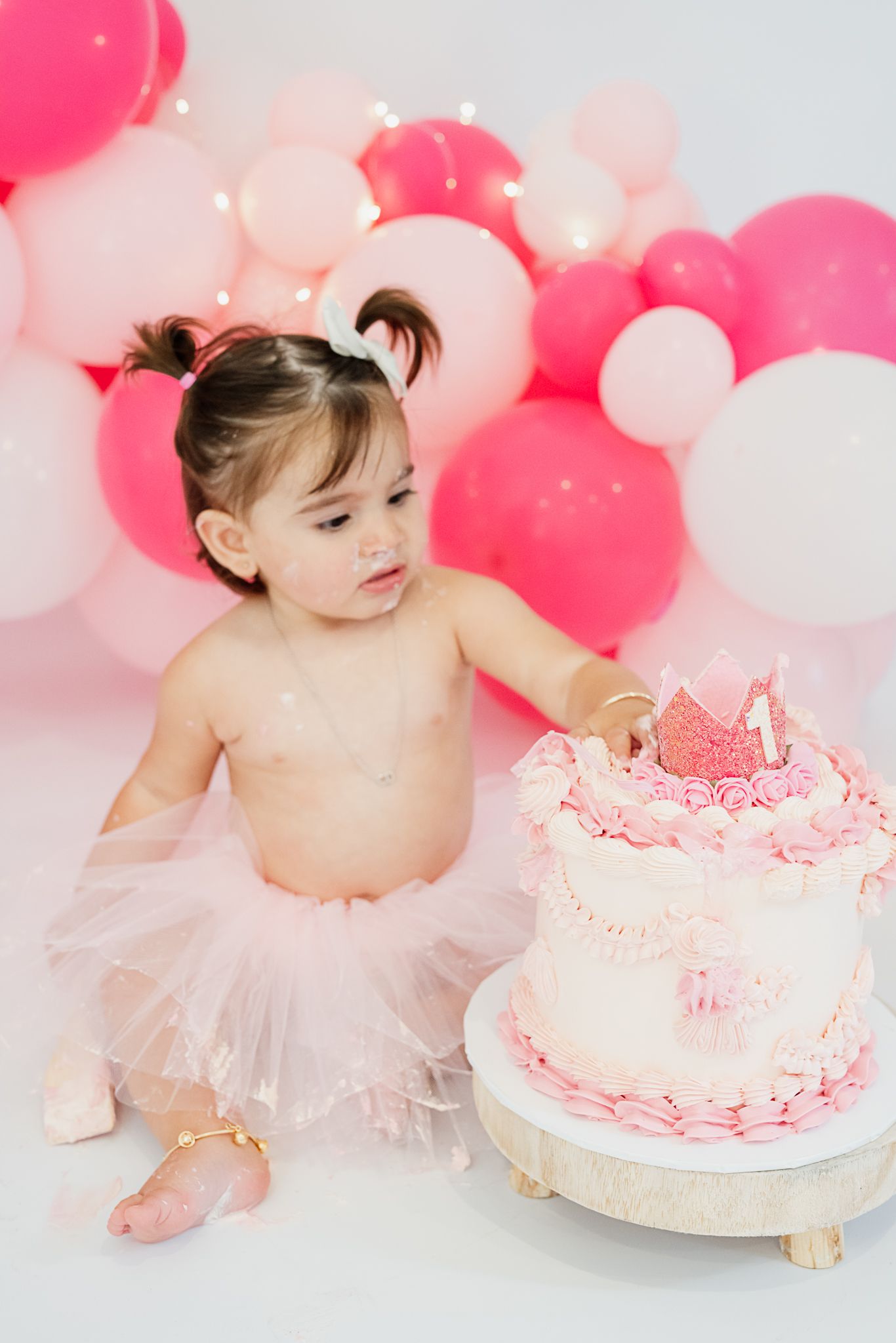 First birthday celebration featuring pink cake and tutu-clad baby against balloon backdrop.