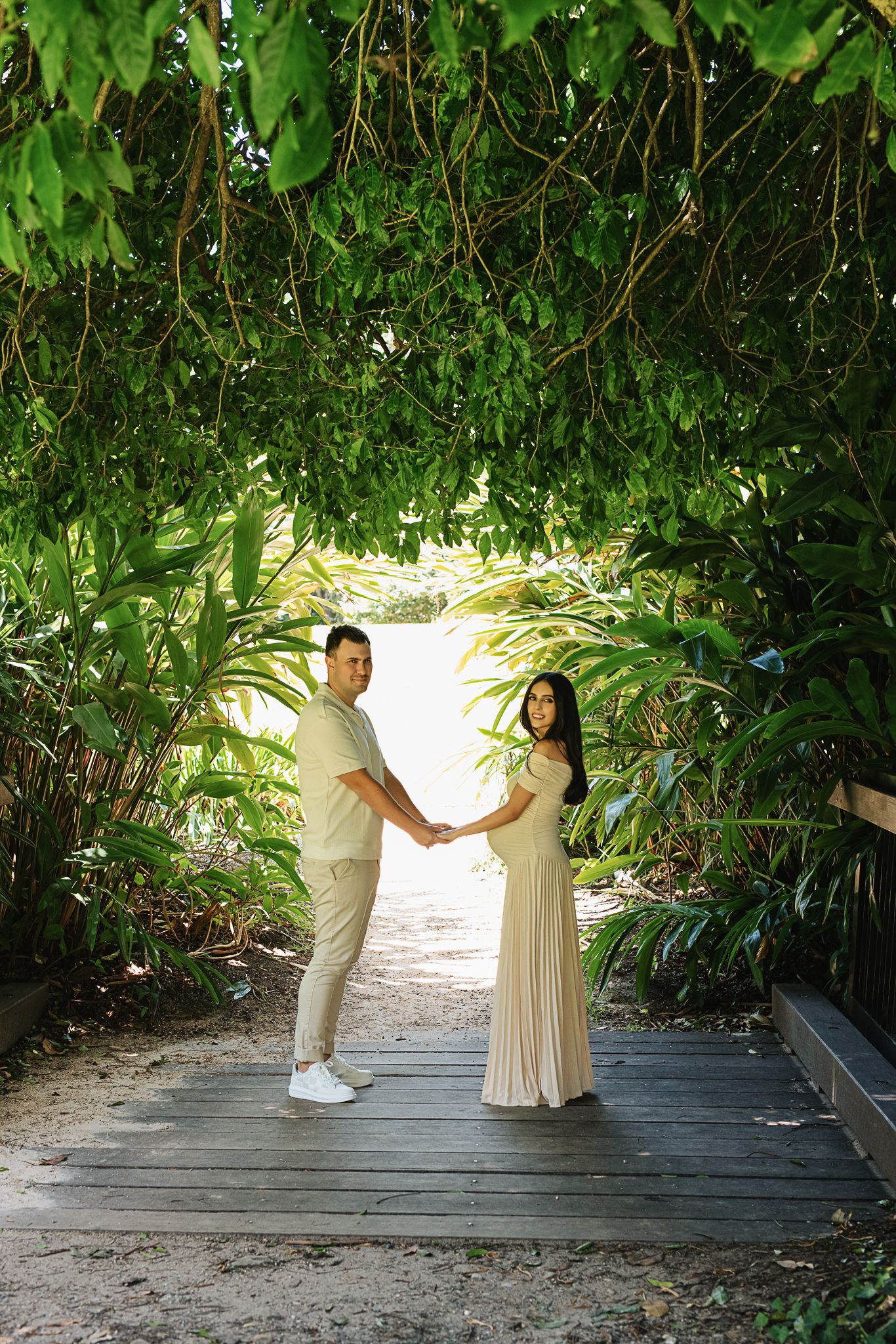 Couple holds hands on wooden path surrounded by lush tropical plants in maternity photo.