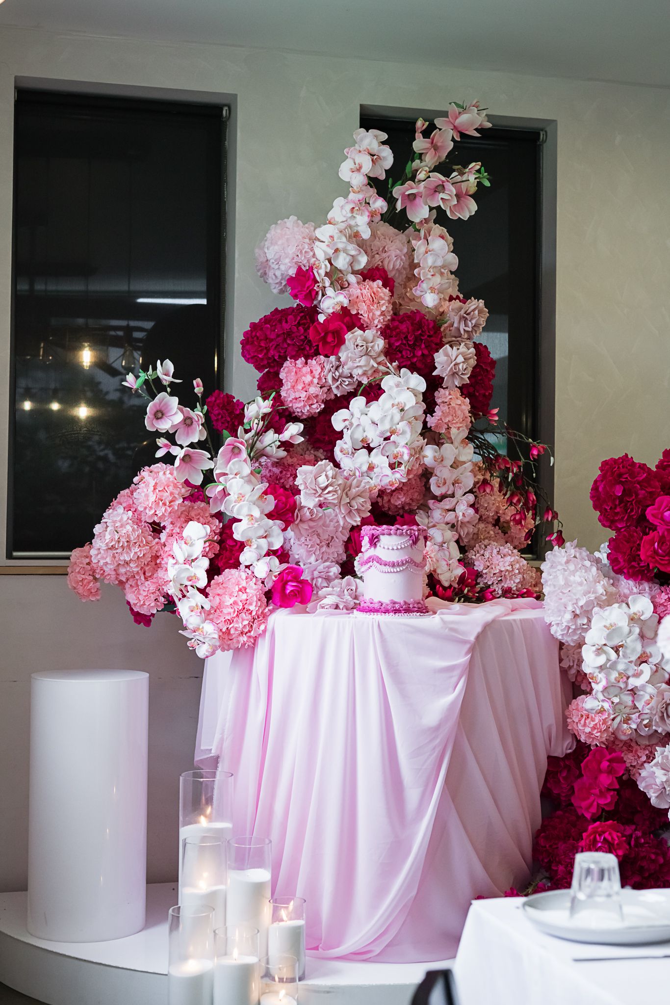 Elegant wedding cake display with cascading pink and white floral arrangements on a pink draped table with glowing candles.
