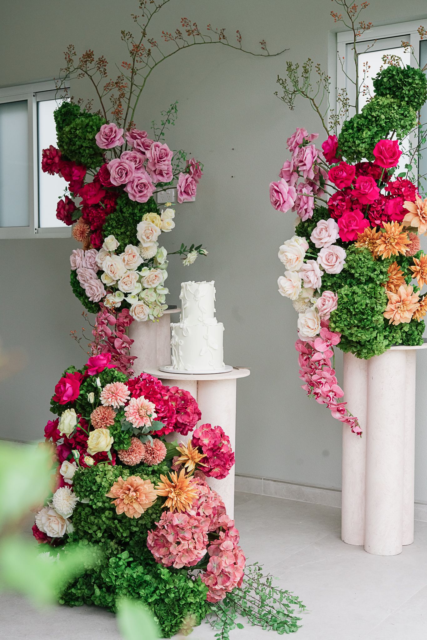 White wedding cake surrounded by lush pink and white floral arrangements on pedestals.
