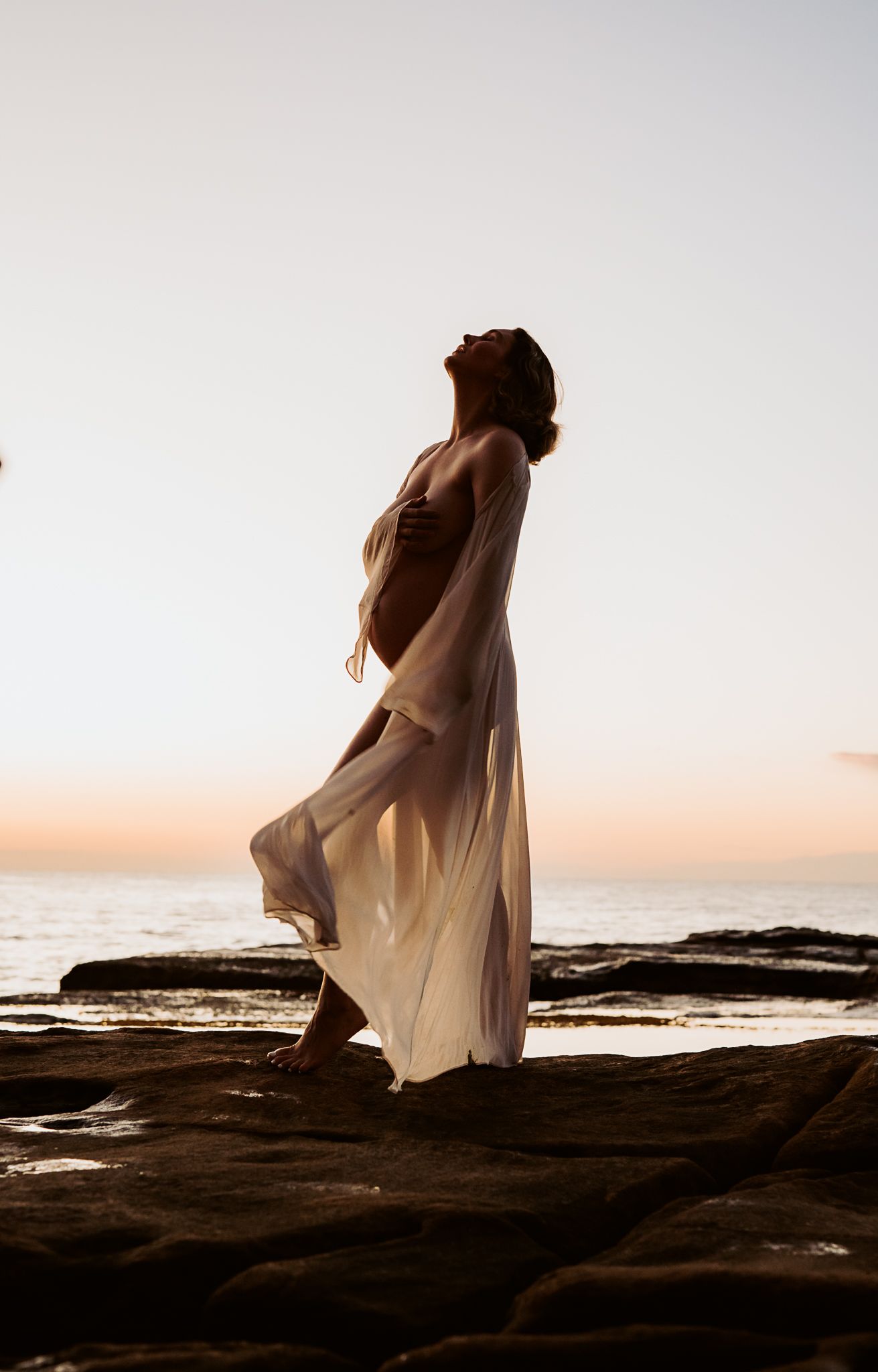 Figure in light dress poses gracefully on coastal rocks during golden hour.