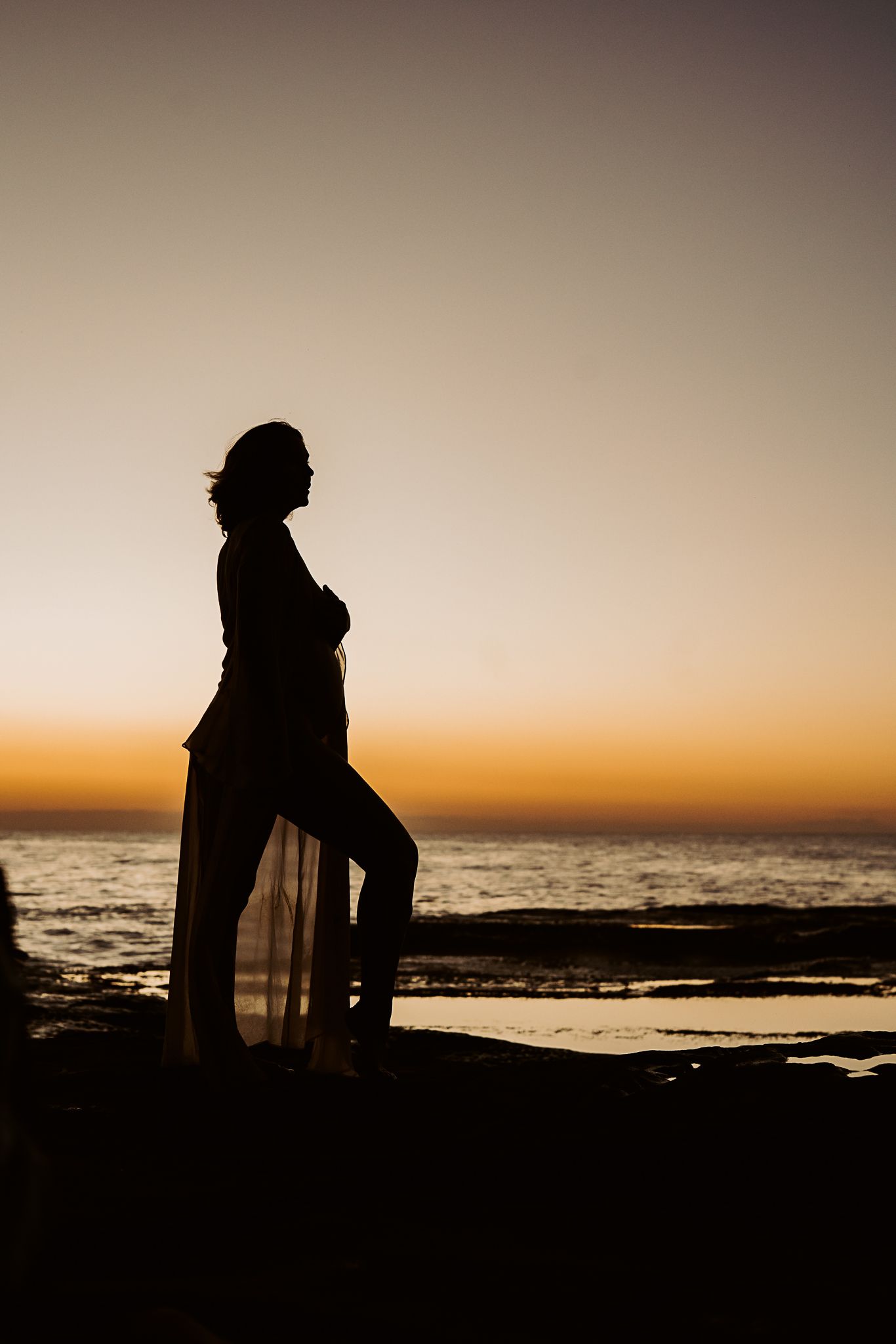 Silhouetted figure stands on rocky coast during golden hour, creating dramatic maternity portrait.